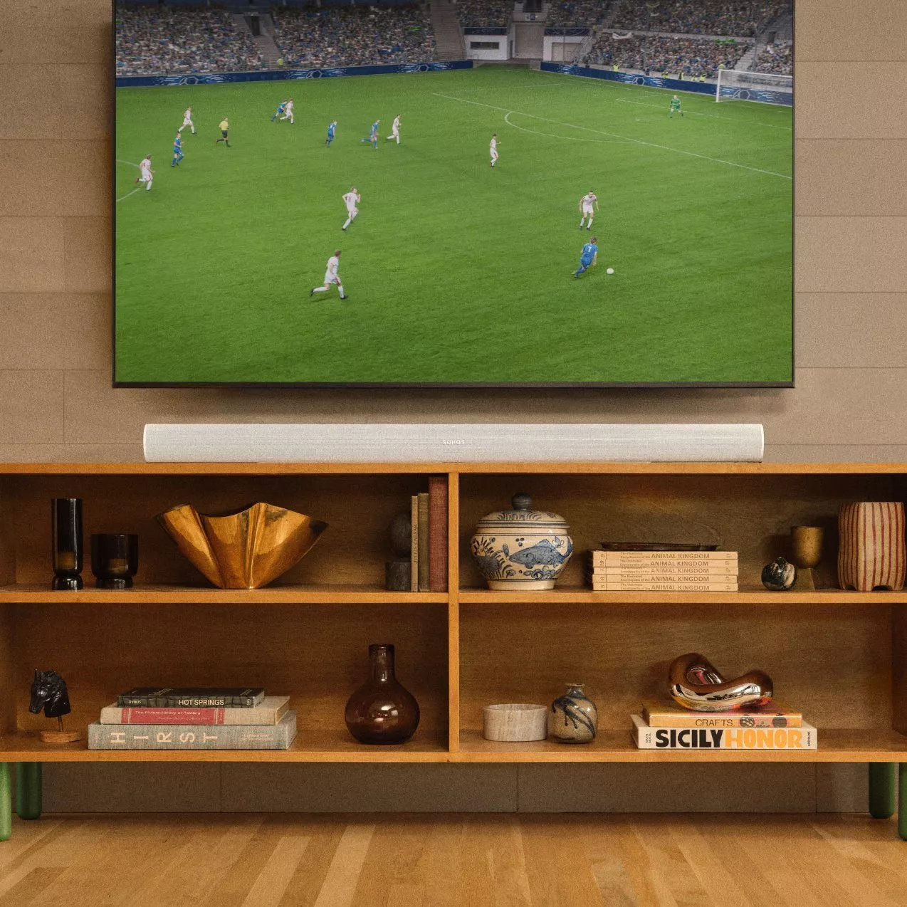 A white Sonos soundbar resting on a wooden media console directly under a wall-mounted TV displaying a soccer match.