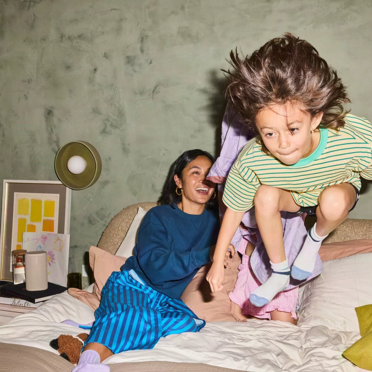 A smiling woman and a child jumping on a bed indoors, with a small light-colored speaker on the nightstand beside them.    JPG