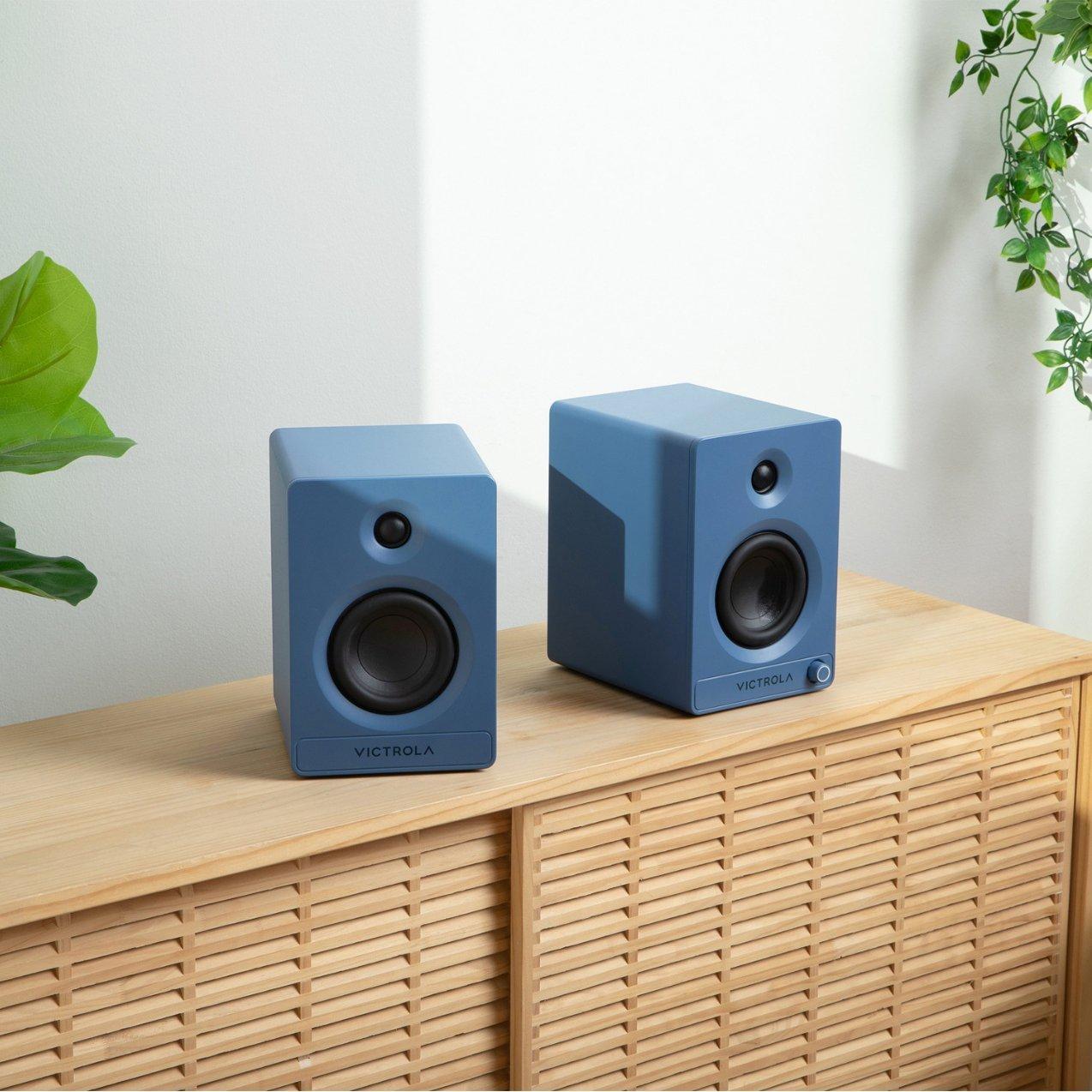 A pair of matte blue Victrola bookshelf speakers sitting side-by-side on a light wood cabinet next to green houseplants in a bright room.