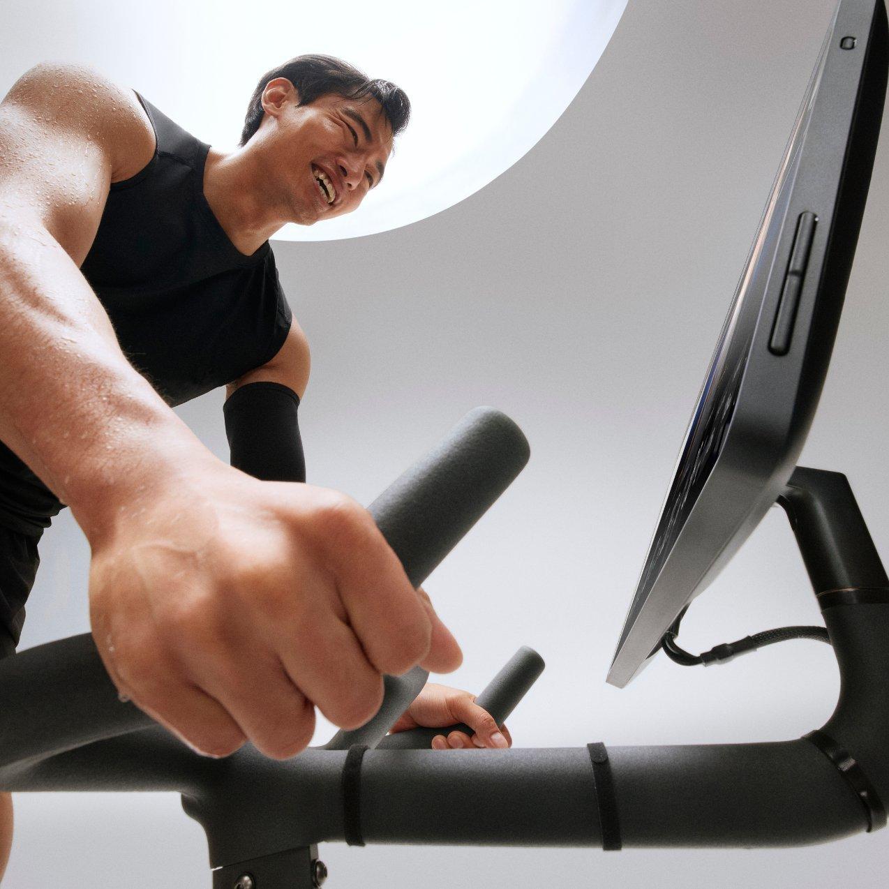 Low-angle shot of a man in a tank top intensely riding a stationary exercise bike while looking at the attached digital screen.