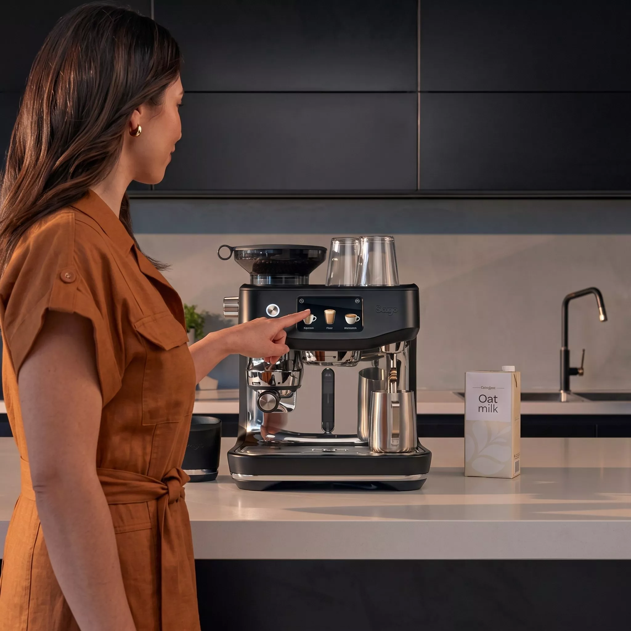 A woman in a brown dress uses the touchscreen on a Sage espresso machine on a kitchen island next to oat milk.