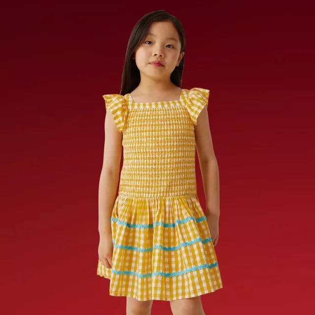 Girl wearing yellow gingham smocked dress with ruffle sleeves and blue trim, studio shot on red background
