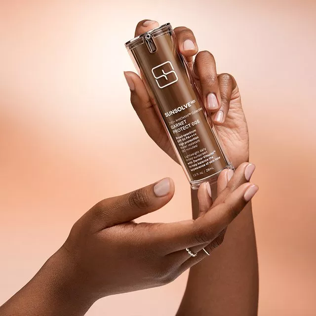 Hands holding Sunsolve MD Garnet Protect serum bottle against peach background, luxury skincare product photo