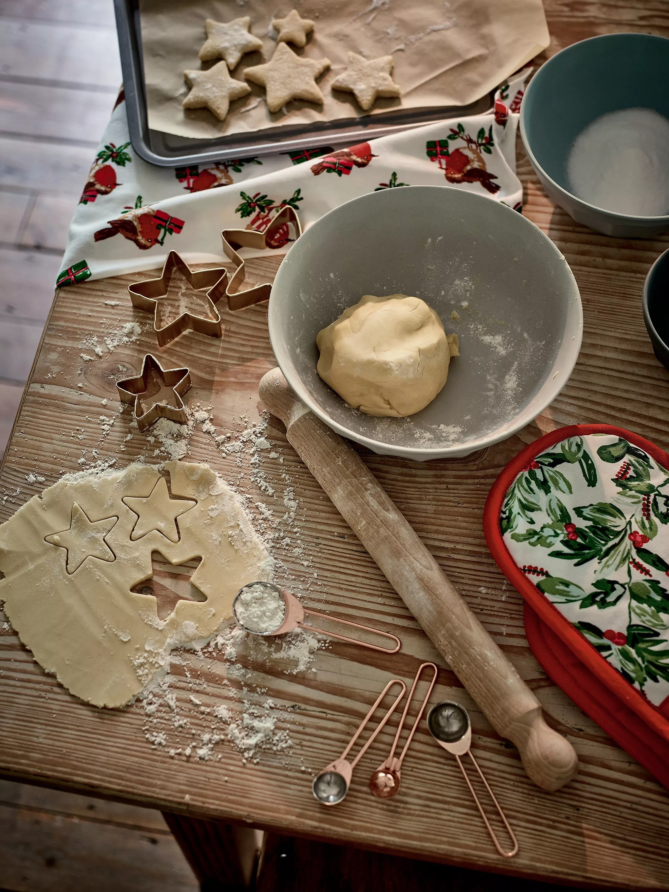 An image of baking with dough in a bowl and star cutters with a rolling pin, oven glove, tea towel and baking try