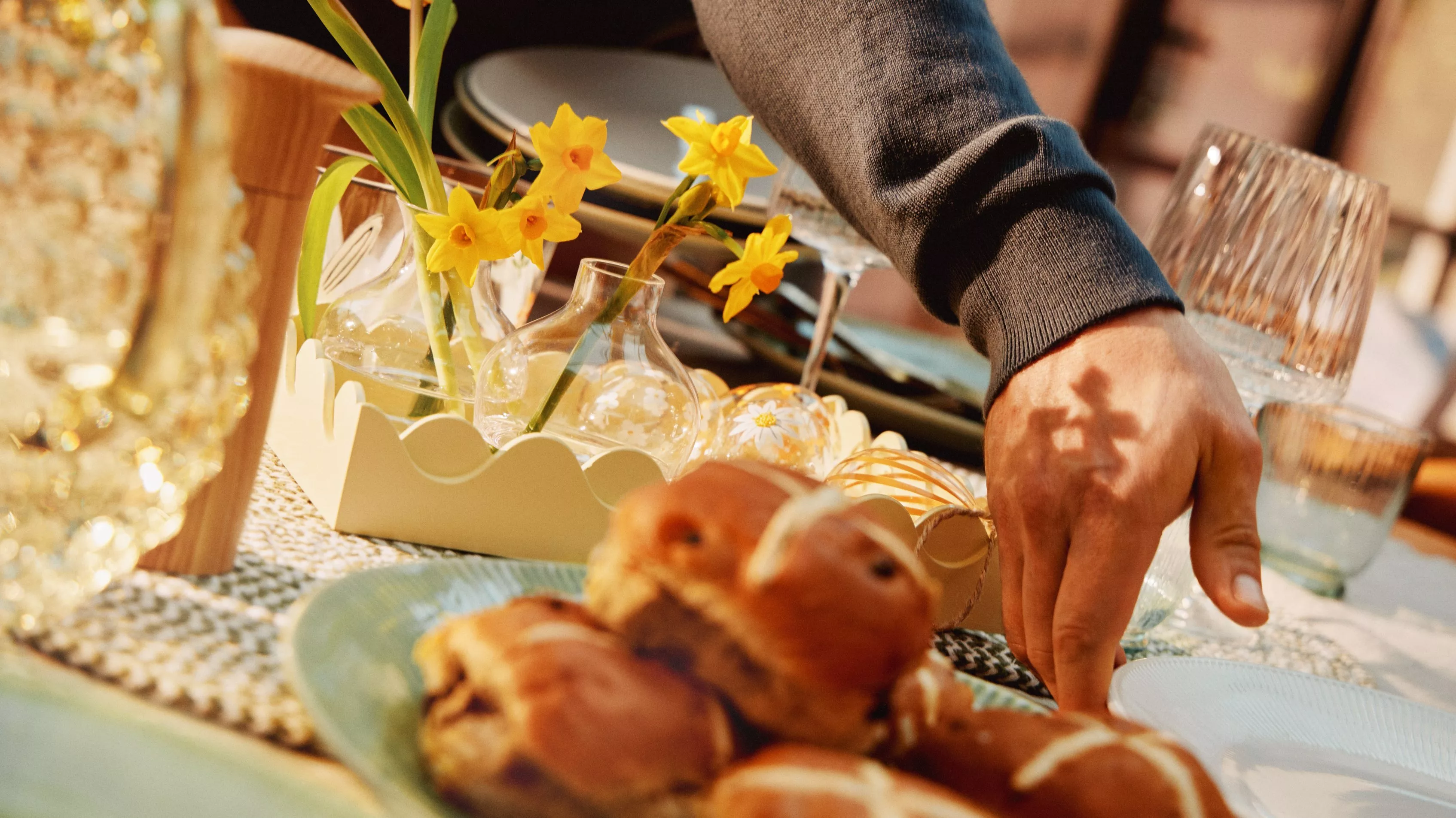A hand reaches for hot cross buns on a table with daffodils and glassware, creating a warm and inviting Easter setting.