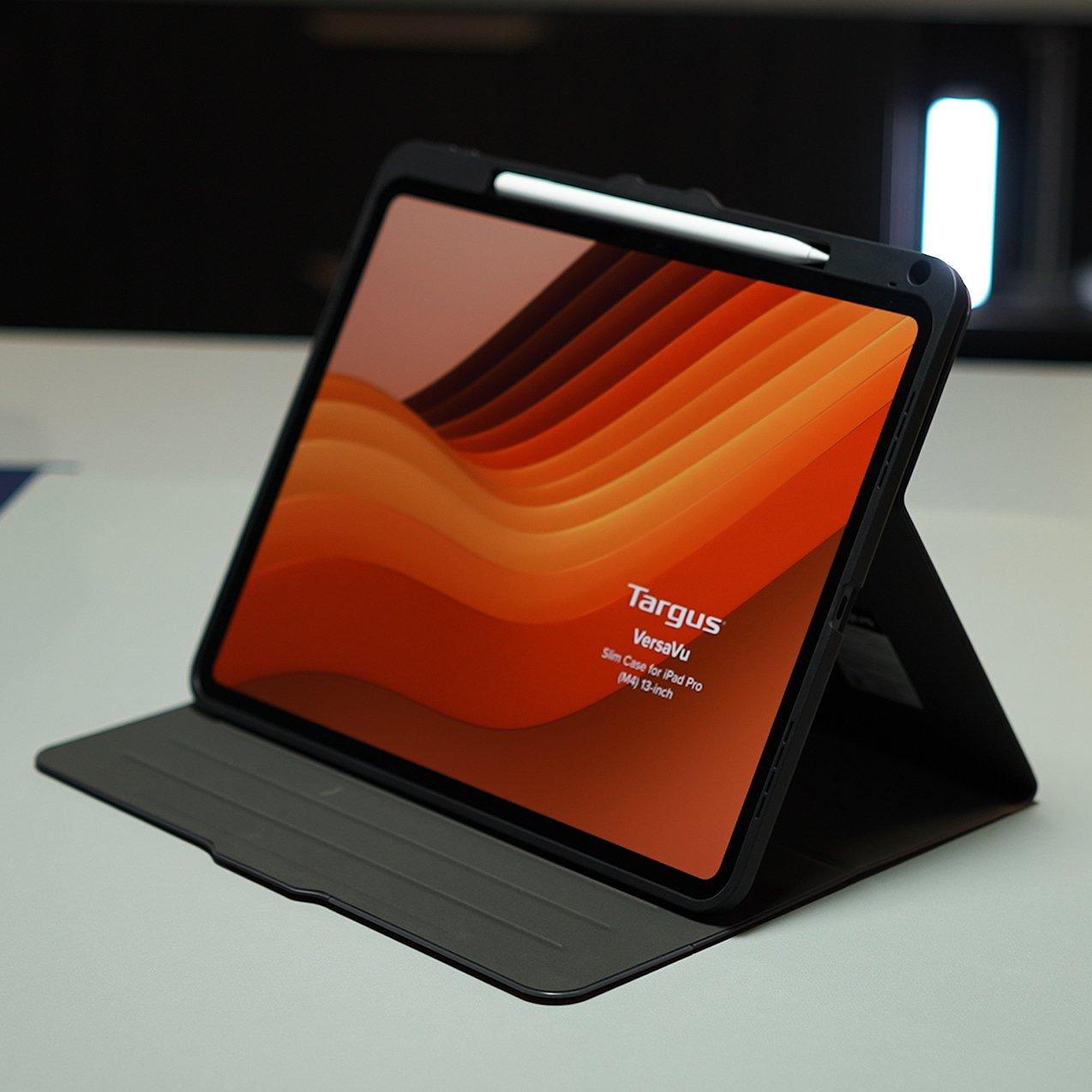 An iPad Pro in a black Targus VersaVu Slim Case set up on a table using its kickstand, featuring an orange abstract wallpaper and a stylus attached at the top.