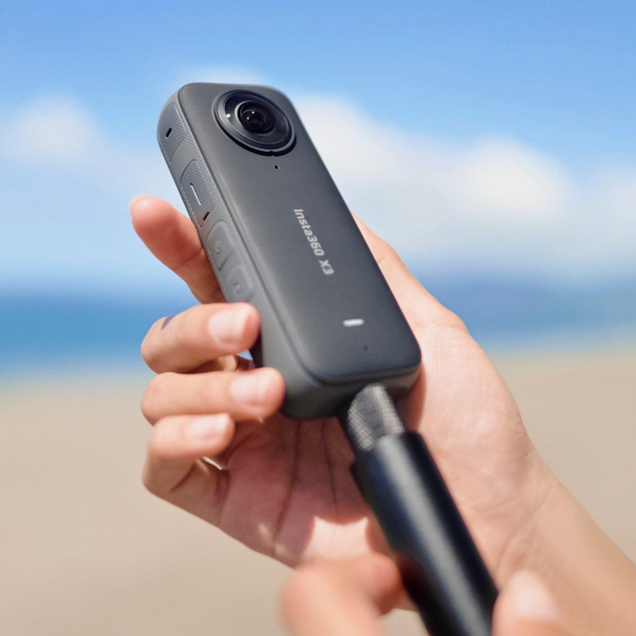 Hand holding Insta360 action camera on selfie stick at beach, blue sky and sea backdrop for travel vlog filming