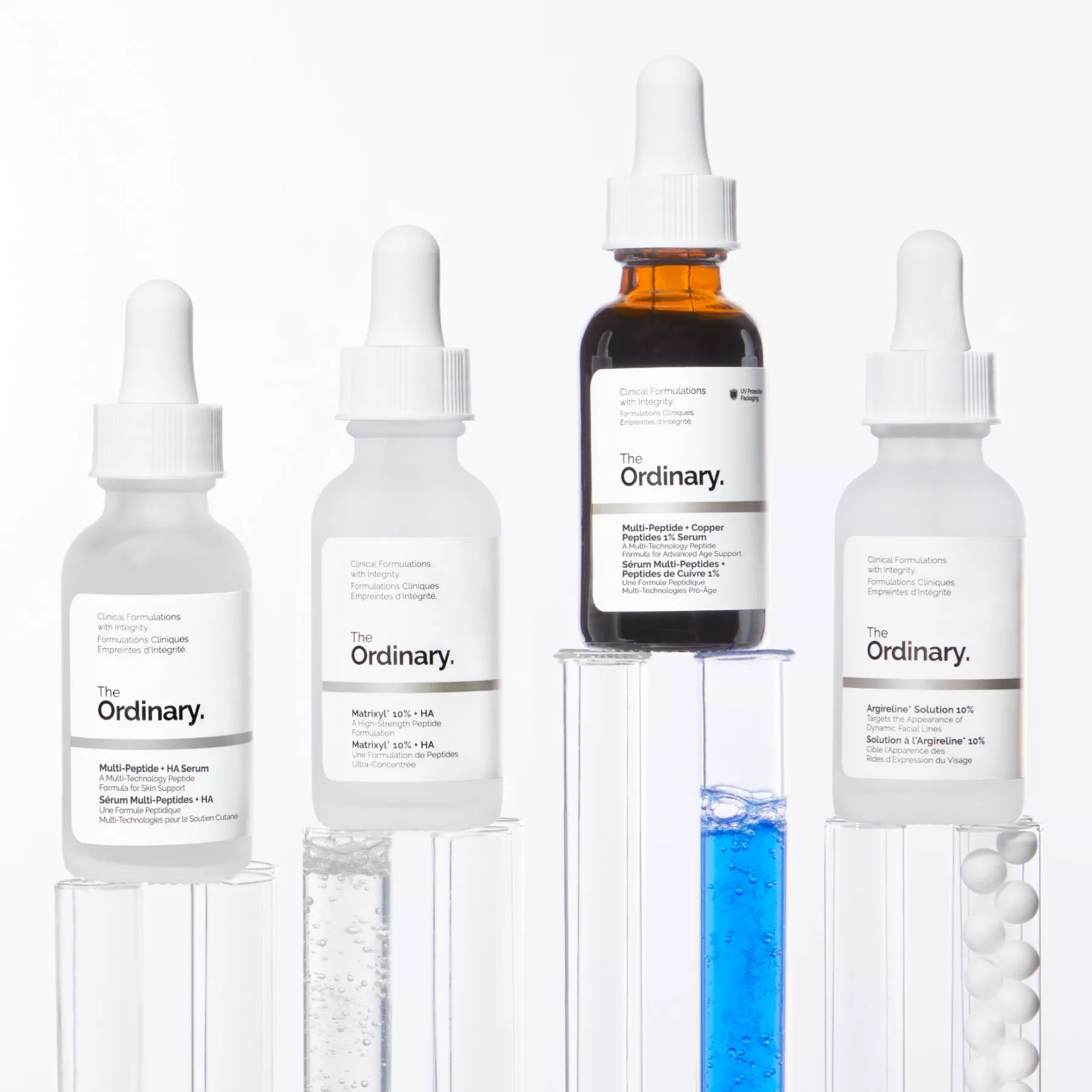 Four The Ordinary serum dropper bottles on clear stands, featuring peptide and copper formulas with blue solution.