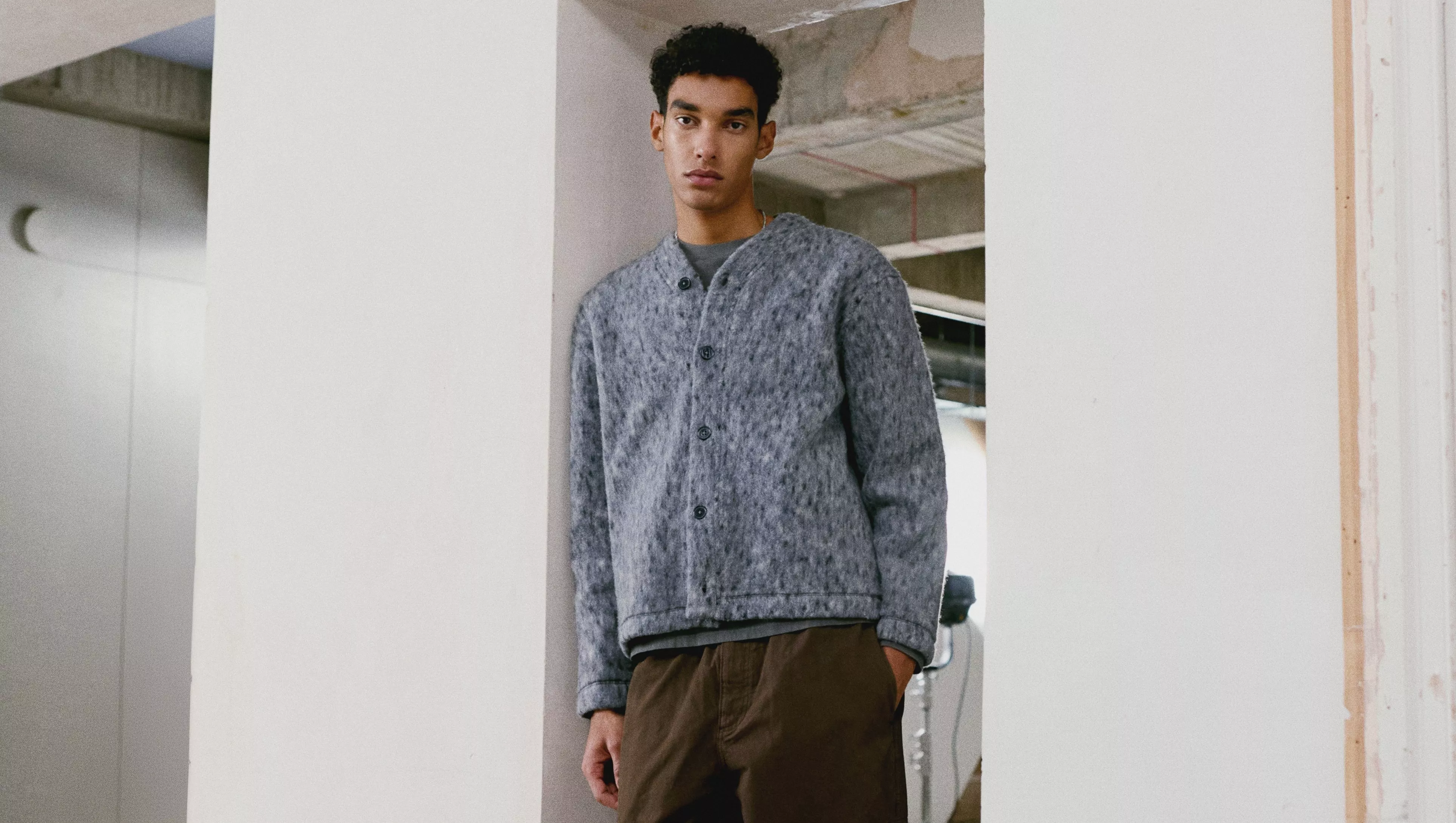 A person wearing a grey buttoned cardigan and brown trousers stands against a white pillar indoors.