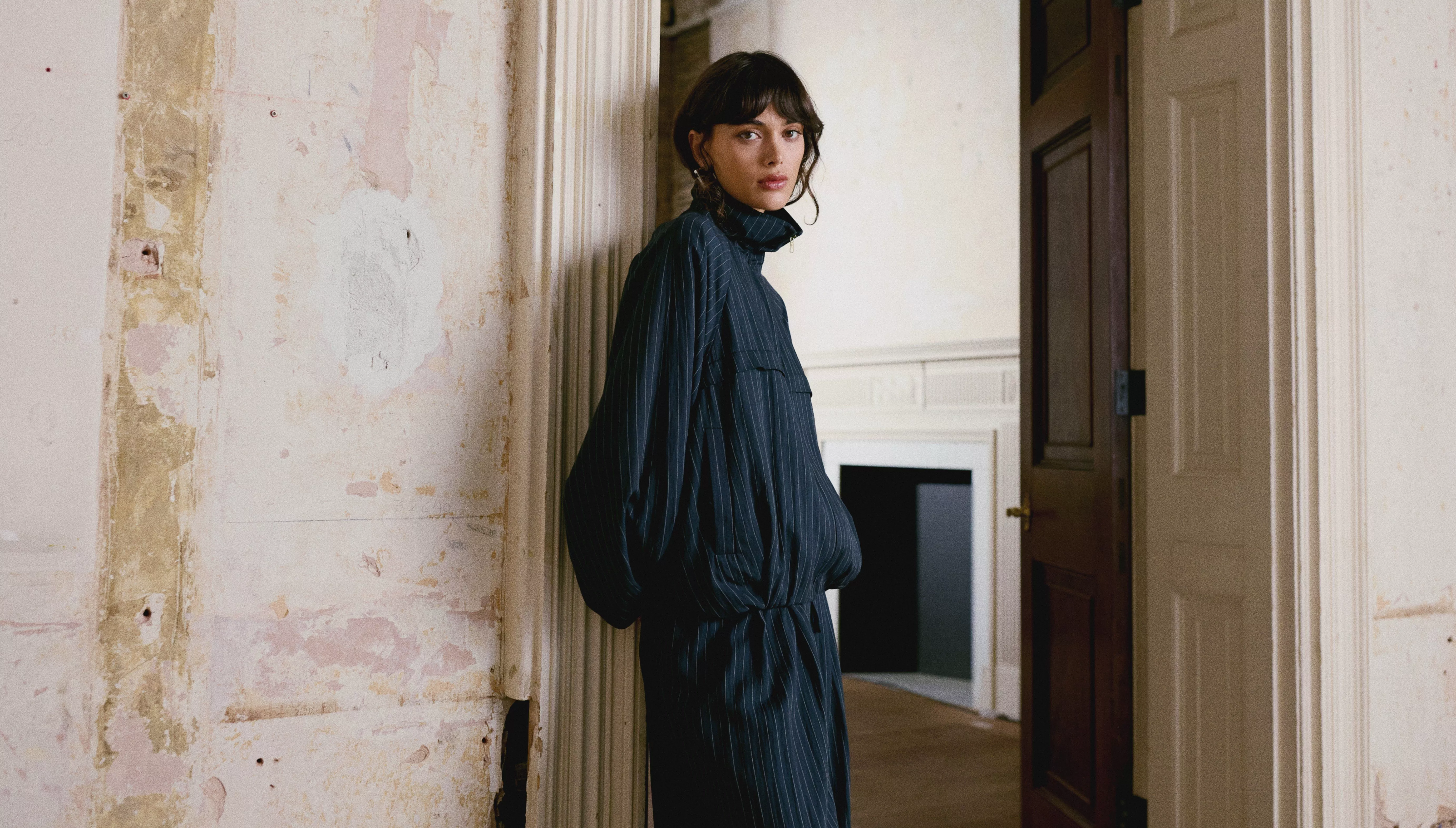 A person in a dark, striped outfit leans against a weathered wall 