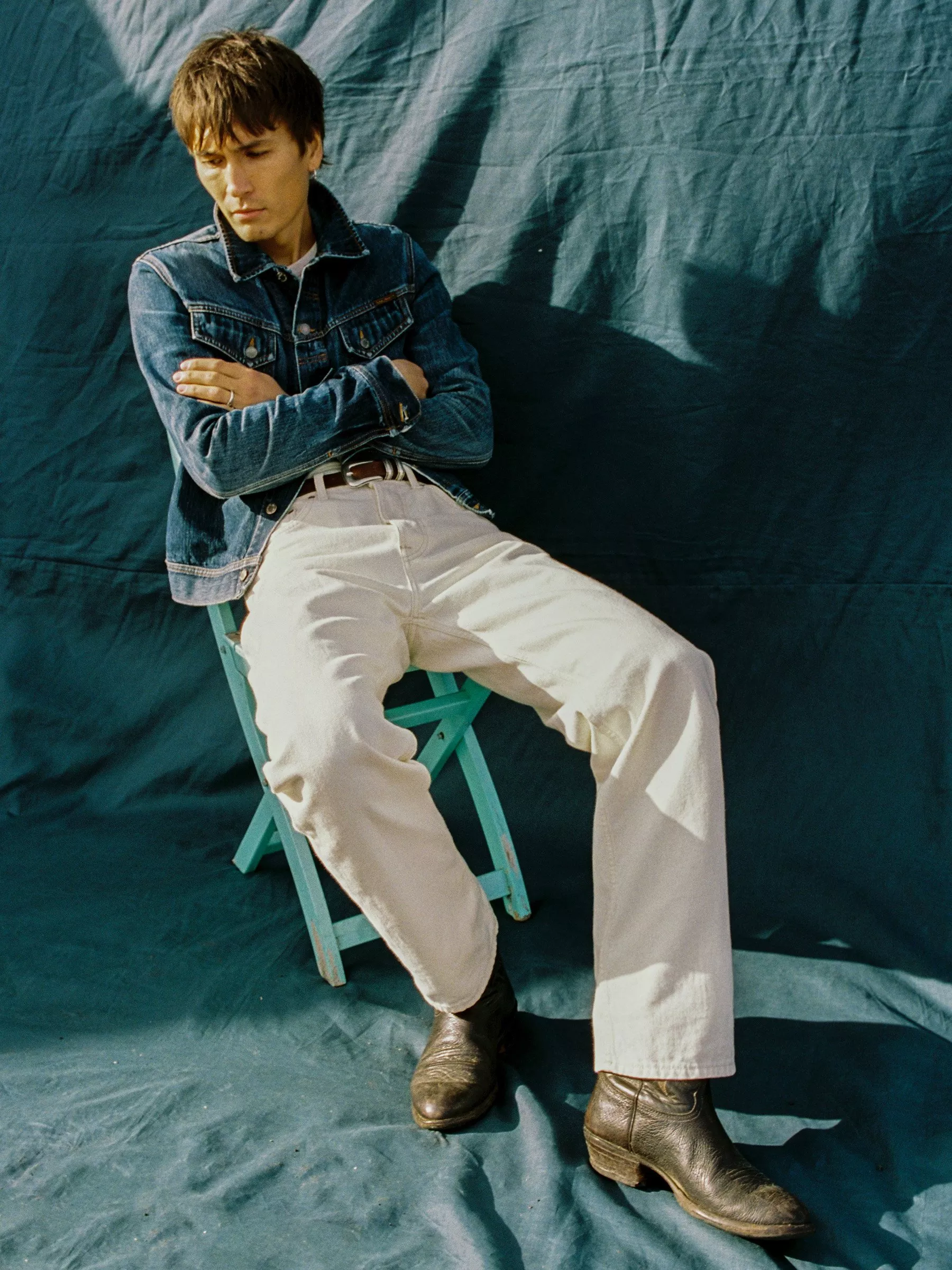 Man in denim jacket and white trousers seated on a teal chair, wearing black boots, against a dark fabric backdrop.
