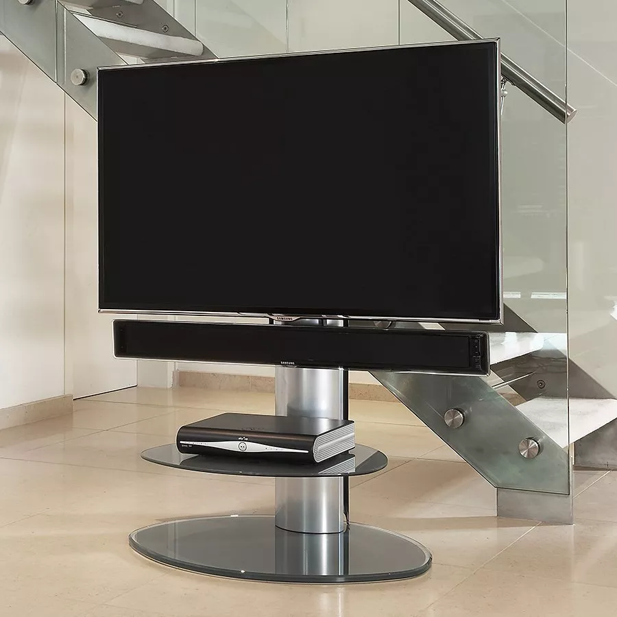 A flat-screen TV with a matching soundbar mounted below it on a silver and glass floor stand. A media box sits on the middle shelf, positioned in front of a modern glass staircase.