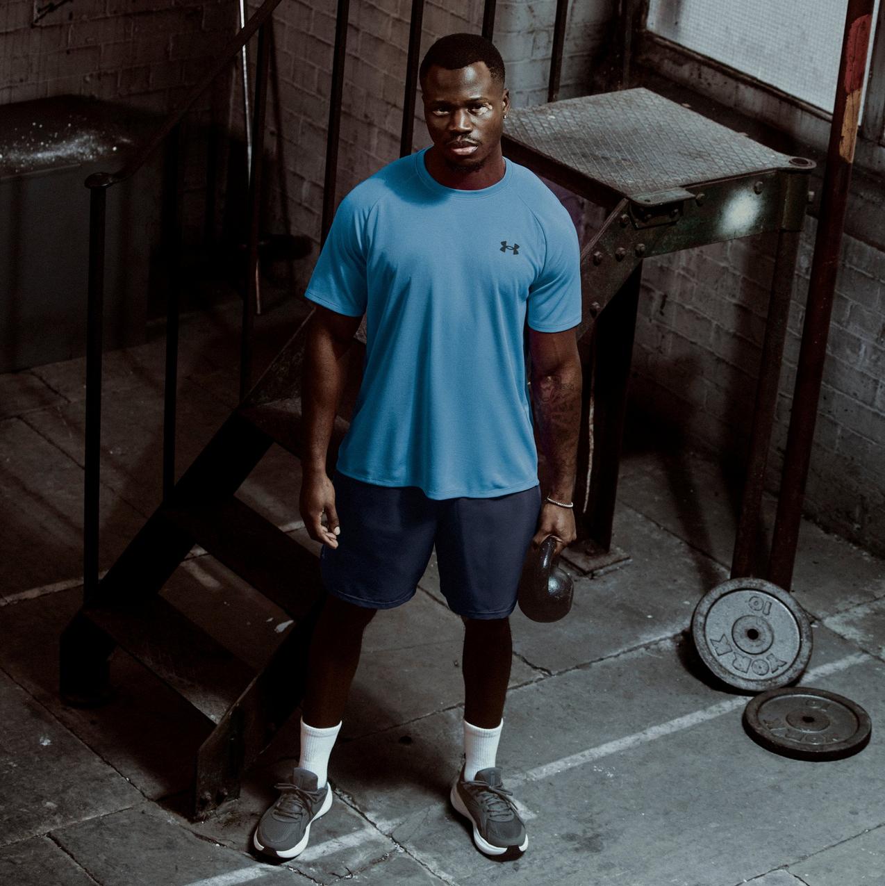 Man holding kettlebell in industrial gym, wearing blue Under Armour training top and shorts, strength workout scene