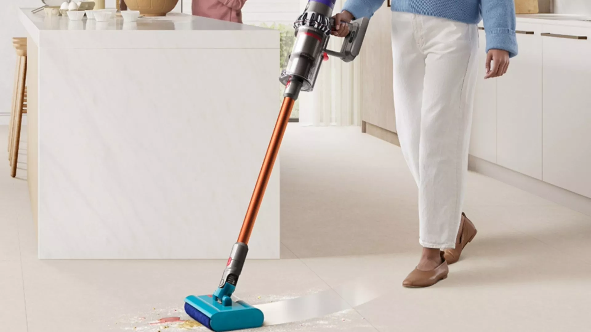 Person using cordless stick vacuum with soft roller head on kitchen floor, cleaning crumbs beside island.