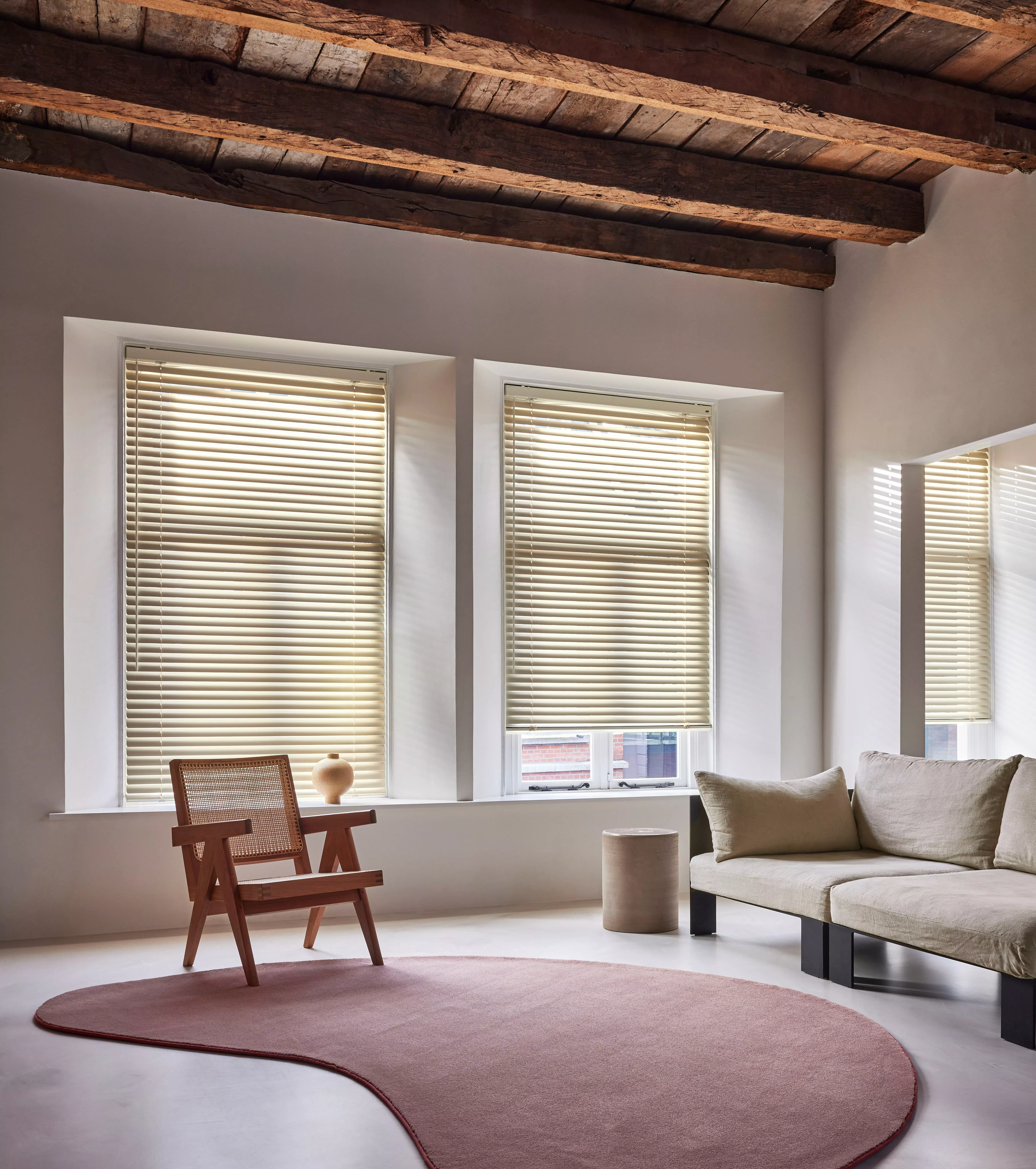 Modern living room with cream Venetian blinds, exposed timber beams, sofa and wooden chair by tall windows