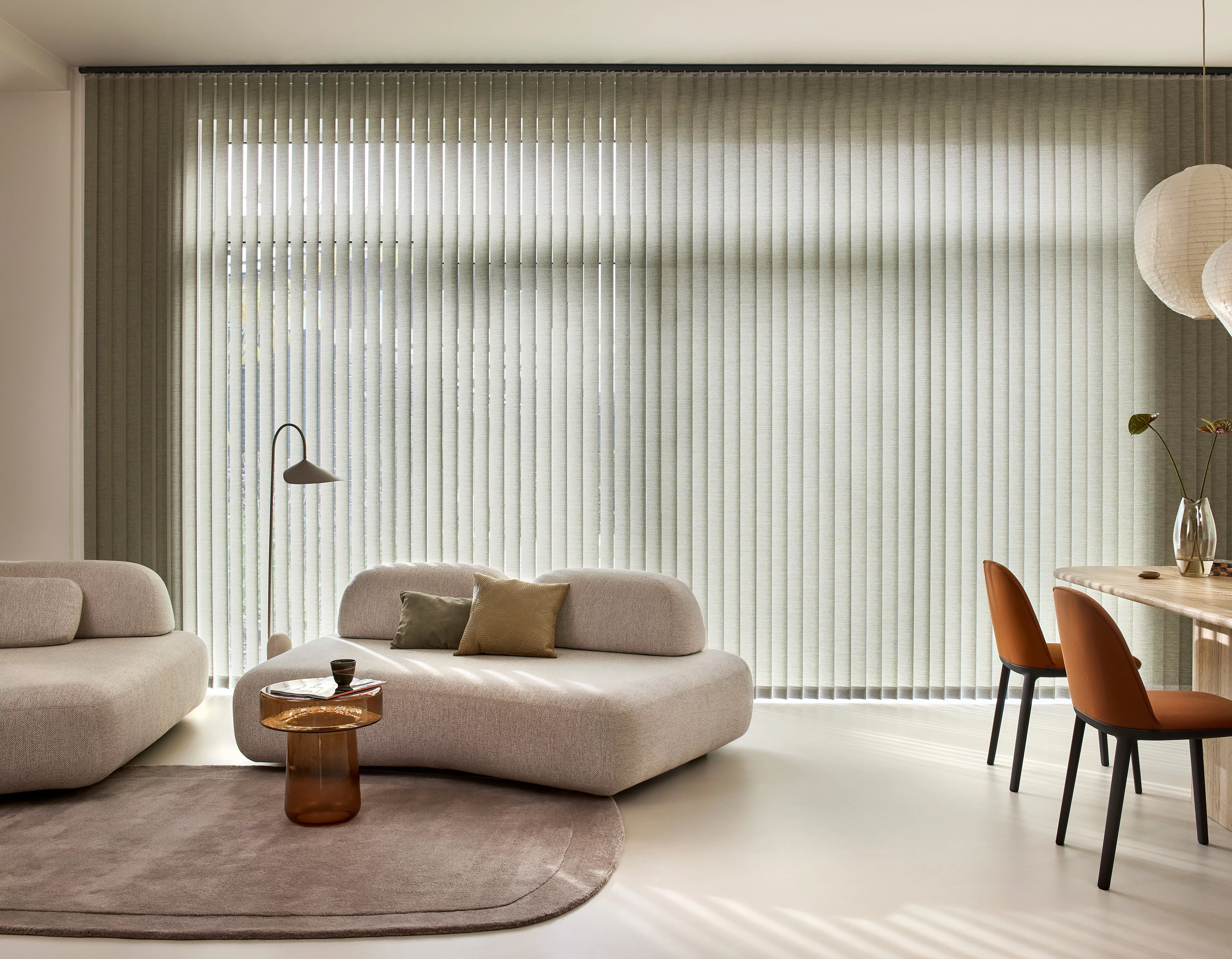 Modern living room with large window vertical blinds, curved grey sofa, glass side table, and dining area chairs
