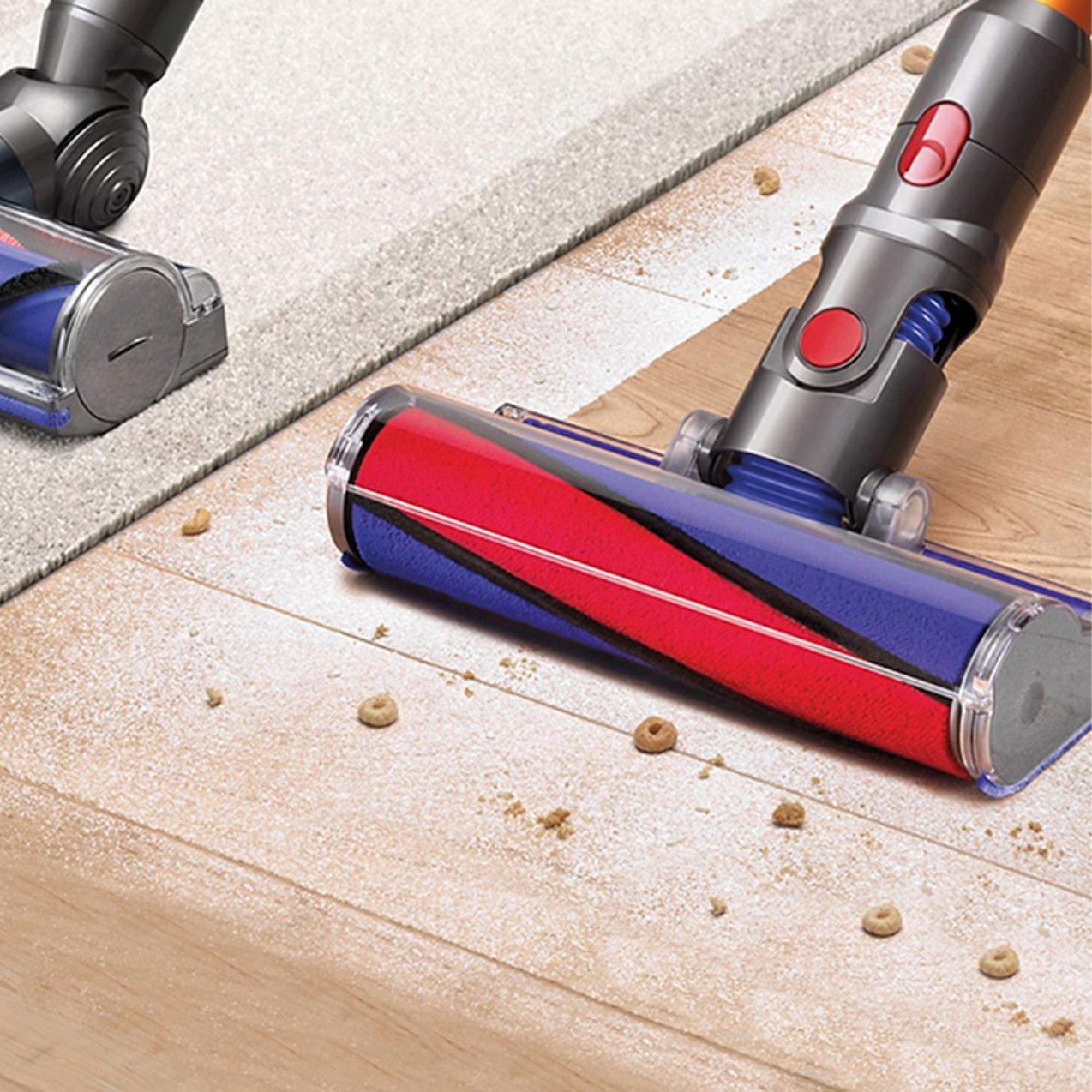 A vacuum cleaner with a red and blue soft roller head cleaning fine dust and cereal pieces off a light wood floor next to a carpeted area.