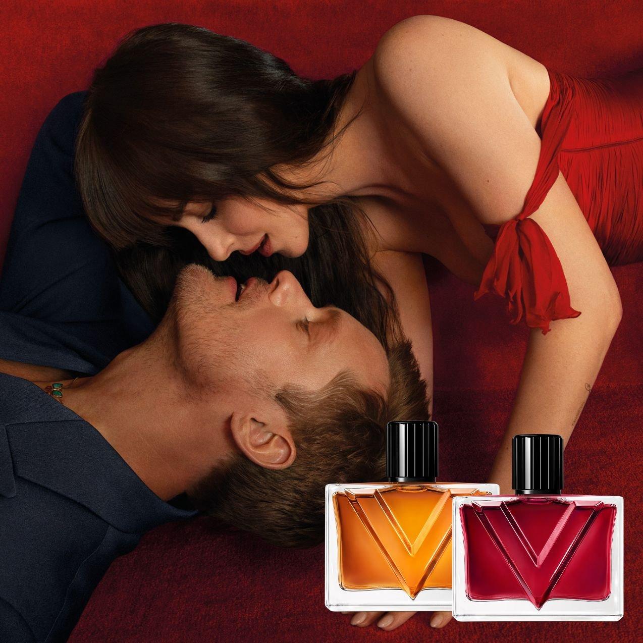 A woman in a red dress leans over a man in a navy suit, faces close together, with two glass perfume bottles featuring a bold "V" design positioned in the foreground.