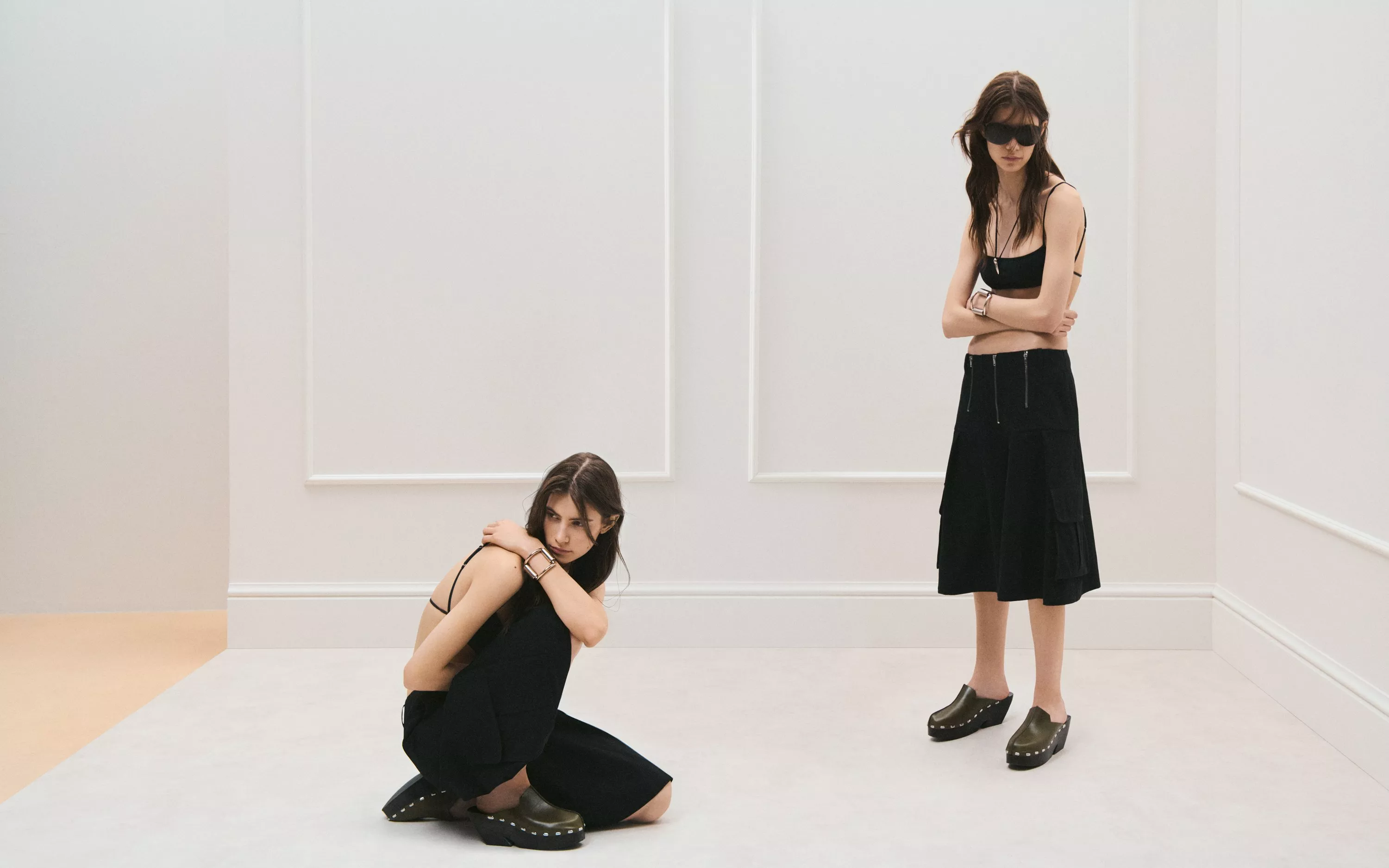 Two fashion models in black outfits and clogs posing in a minimalist white studio for editorial style shoot