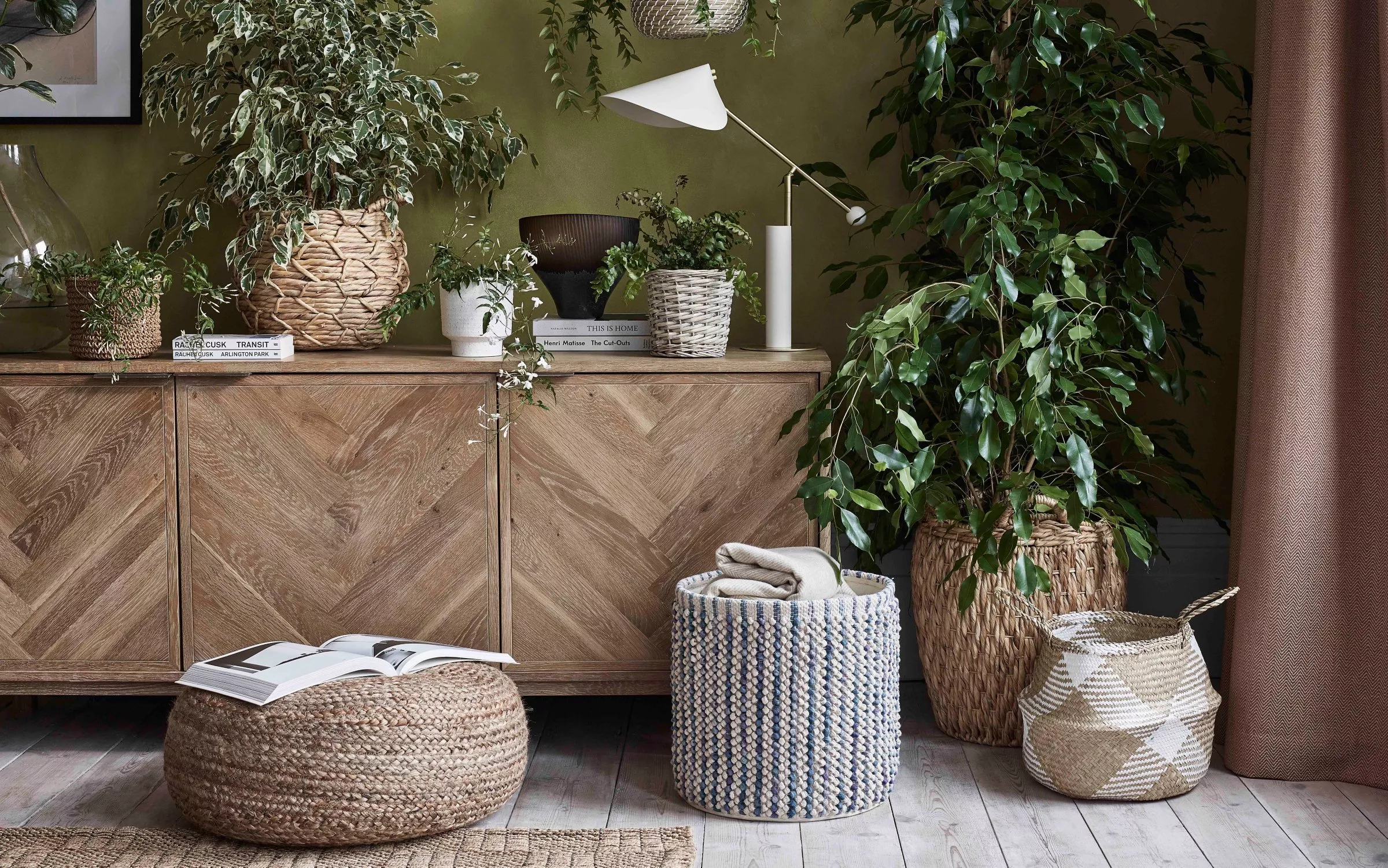 Indoor plants on wooden sideboard with woven baskets, pouffes and modern lamp in cosy living room decor