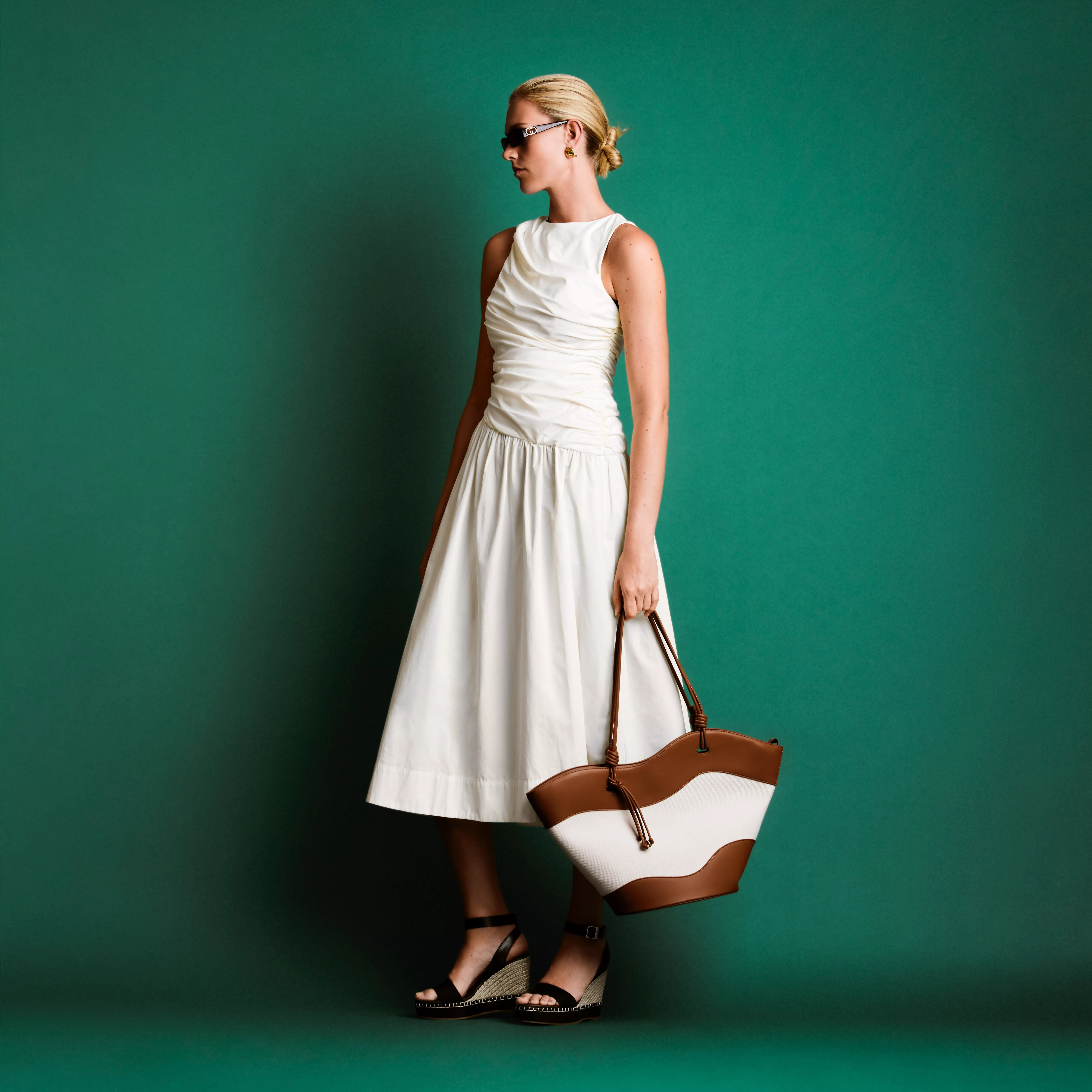 Woman in white midi dress holding brown and cream tote bag, standing against teal studio backdrop, fashion shot