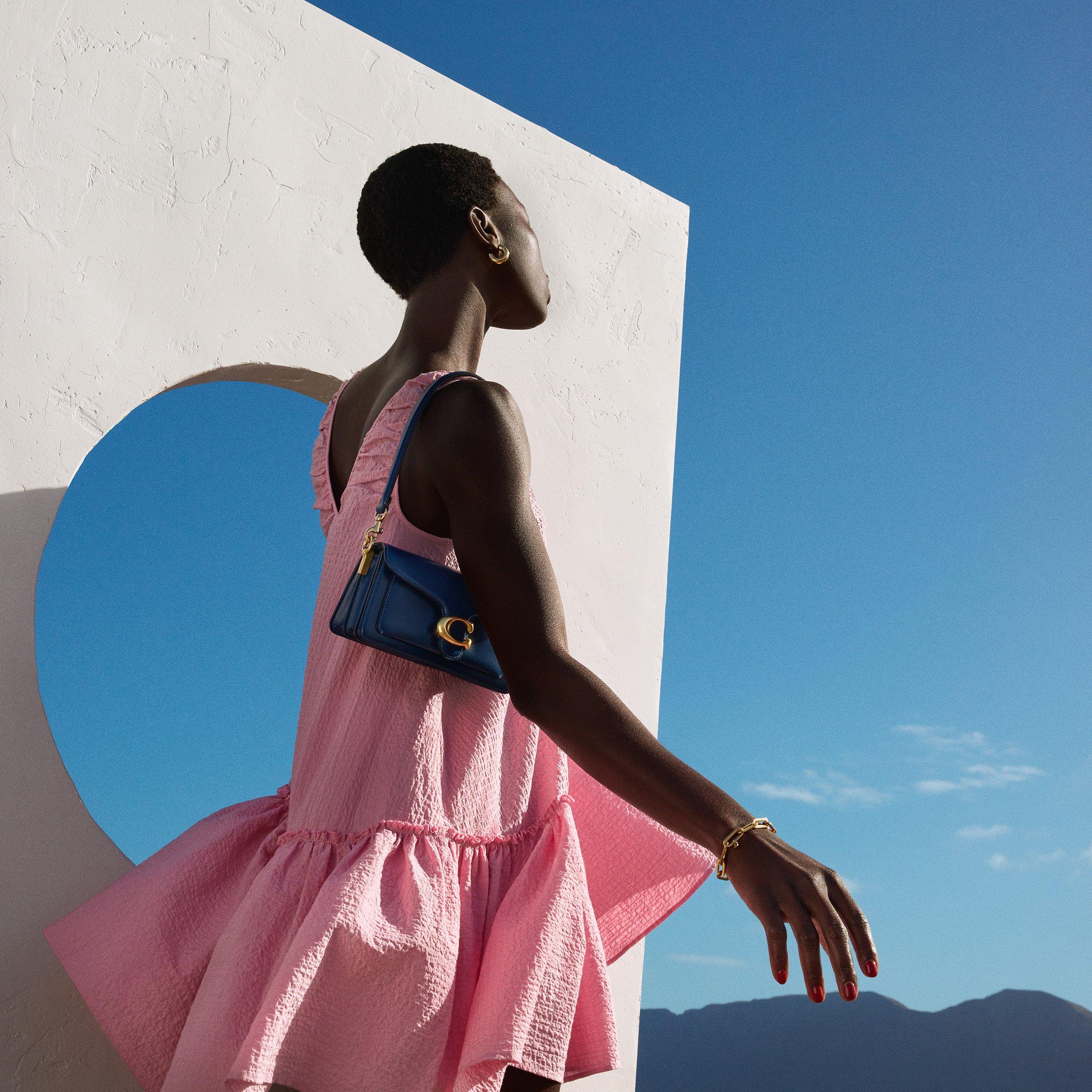 Woman in a pink textured dress poses outdoors with a blue shoulder bag, gold jewelry, and a modern white backdrop against a clear blue sky.