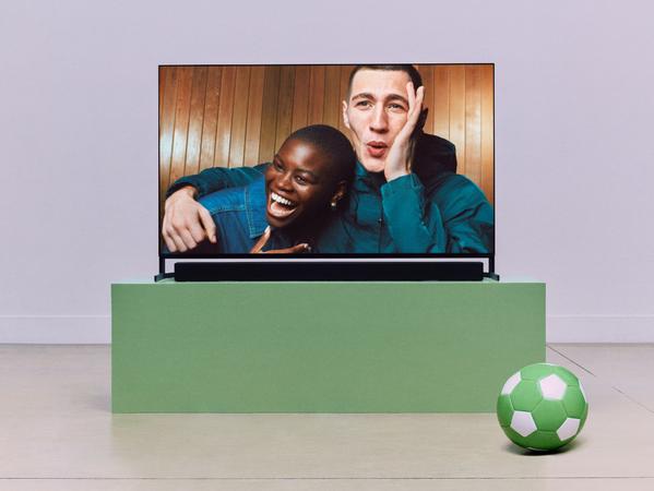 Large flat-screen TV with soundbar showing friends laughing, with a green football on the floor in front