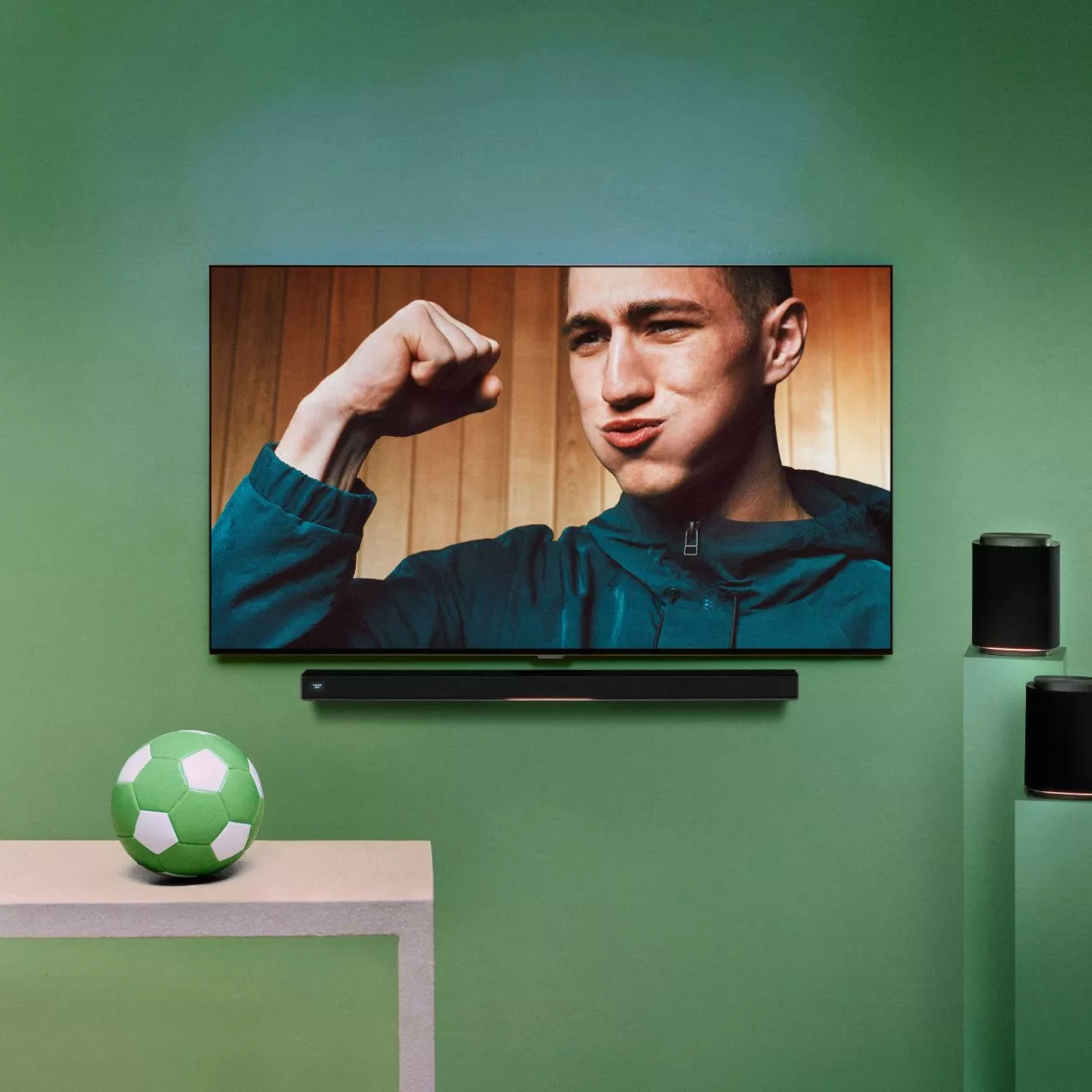 Wall-mounted TV showing football fan celebrating, with soundbar, speakers and green football in modern living room
