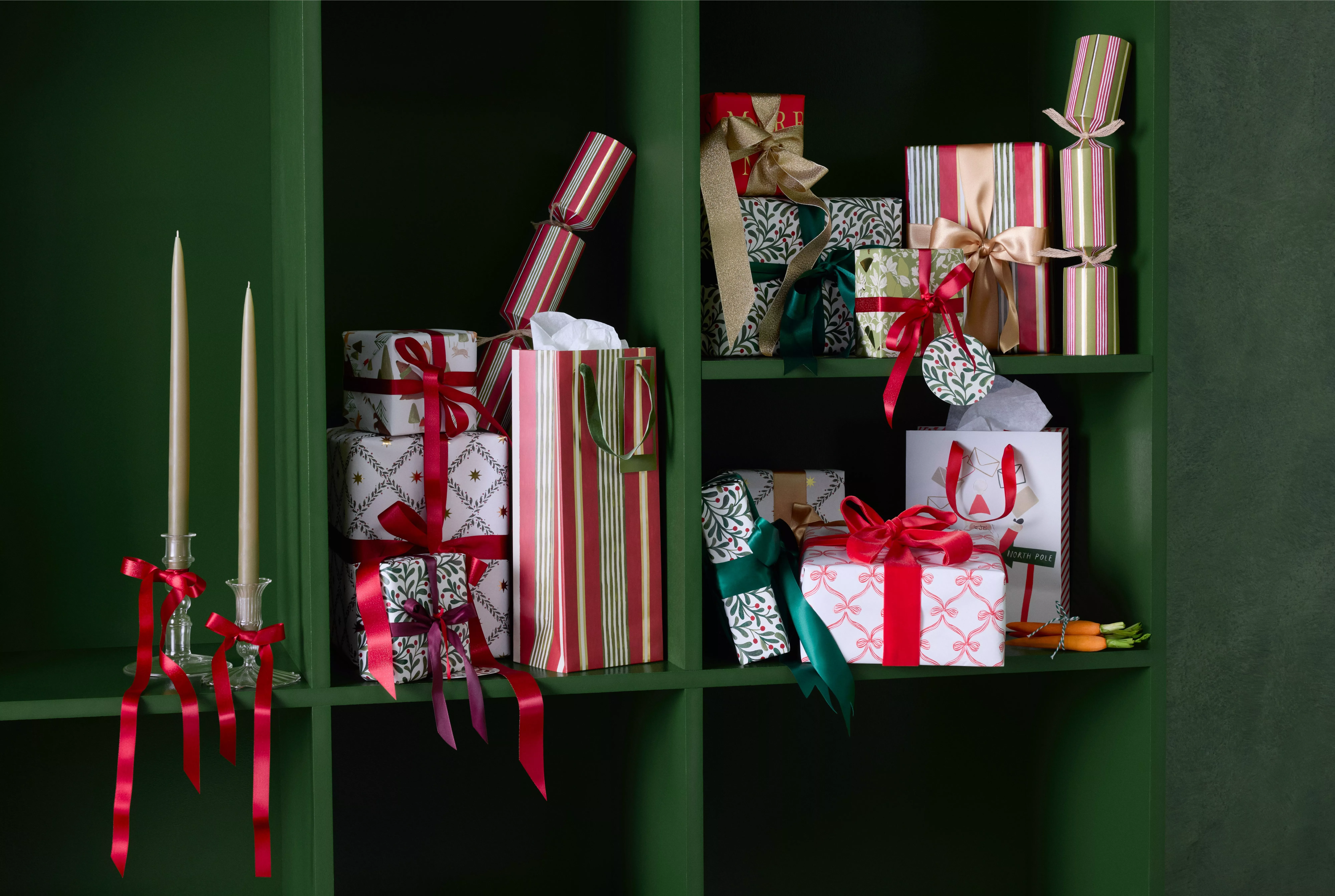 Festive Christmas gift boxes with red ribbons on green shelving, with taper candles and wrapped presents.