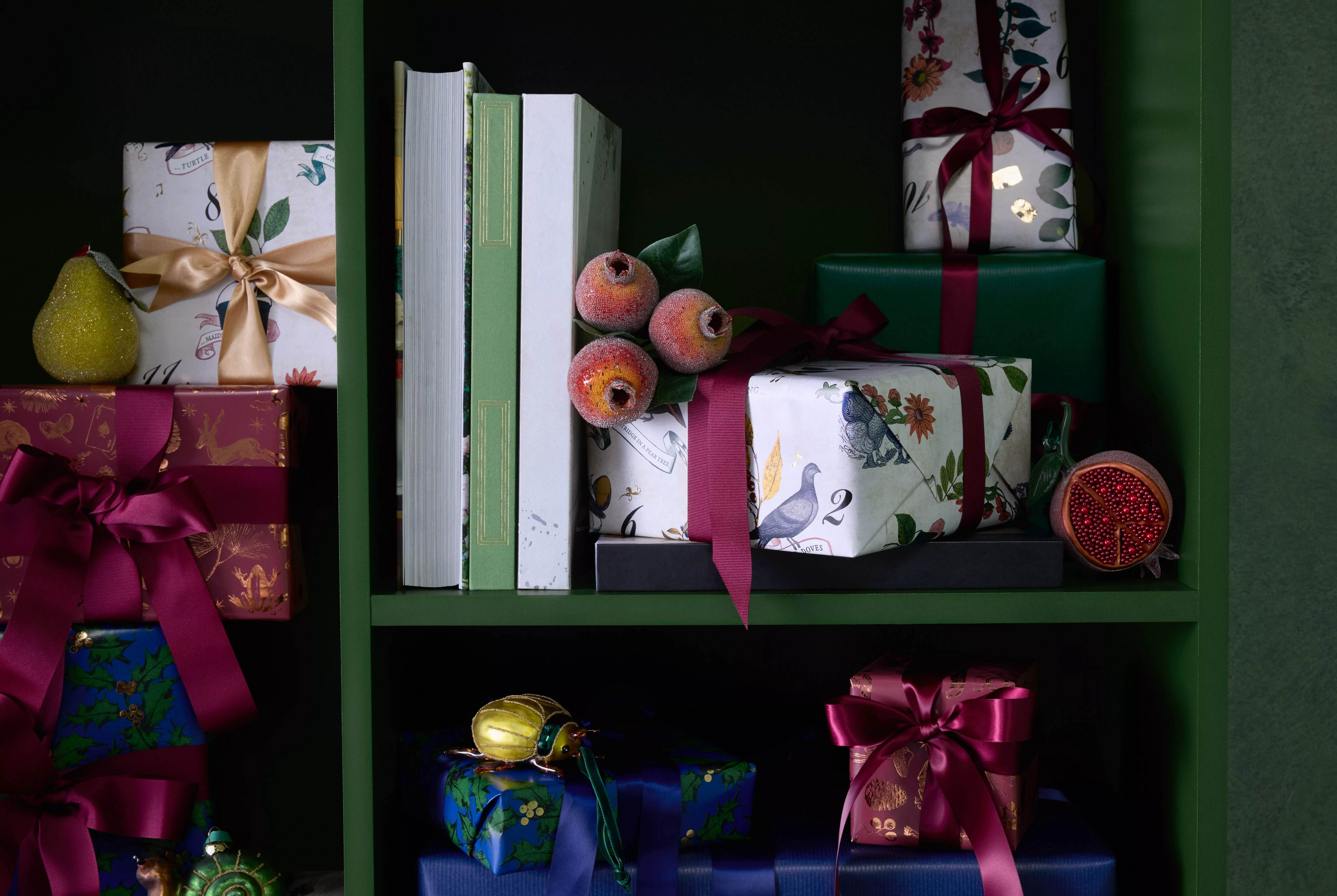 Festive gift boxes with ribbons on a green shelf, beside books, frosted fruit and a pomegranate decoration.