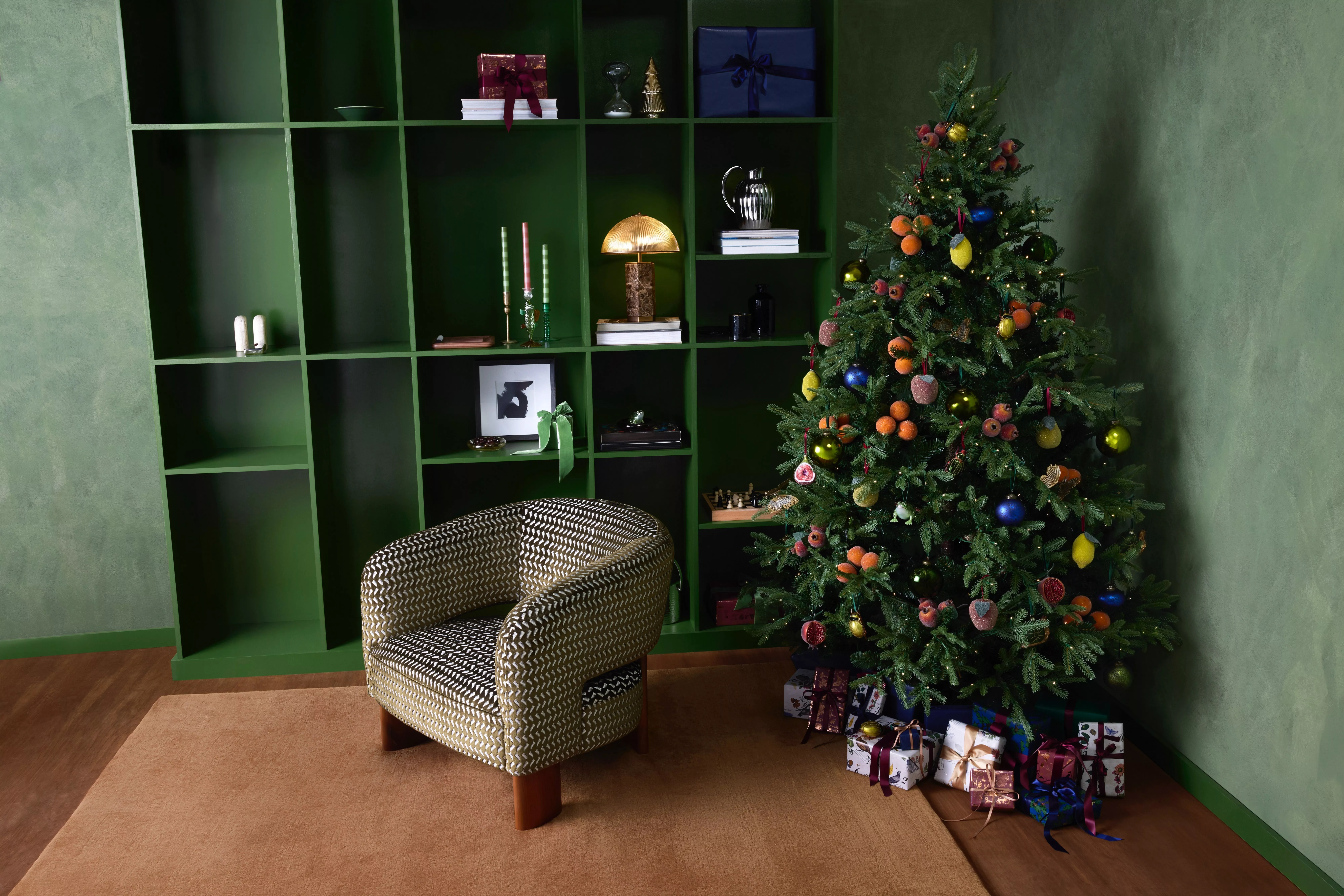Festive living room with decorated Christmas tree, presents, green shelving unit and patterned armchair interior.