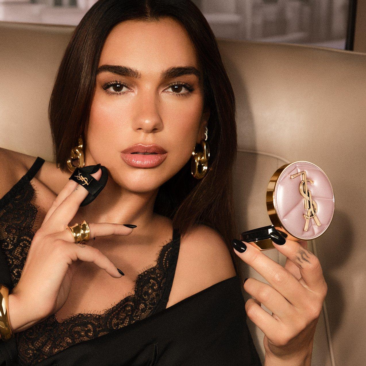 Dua Lipa holds a pink and gold YSL cushion foundation compact while pressing a black applicator pad to her cheek.