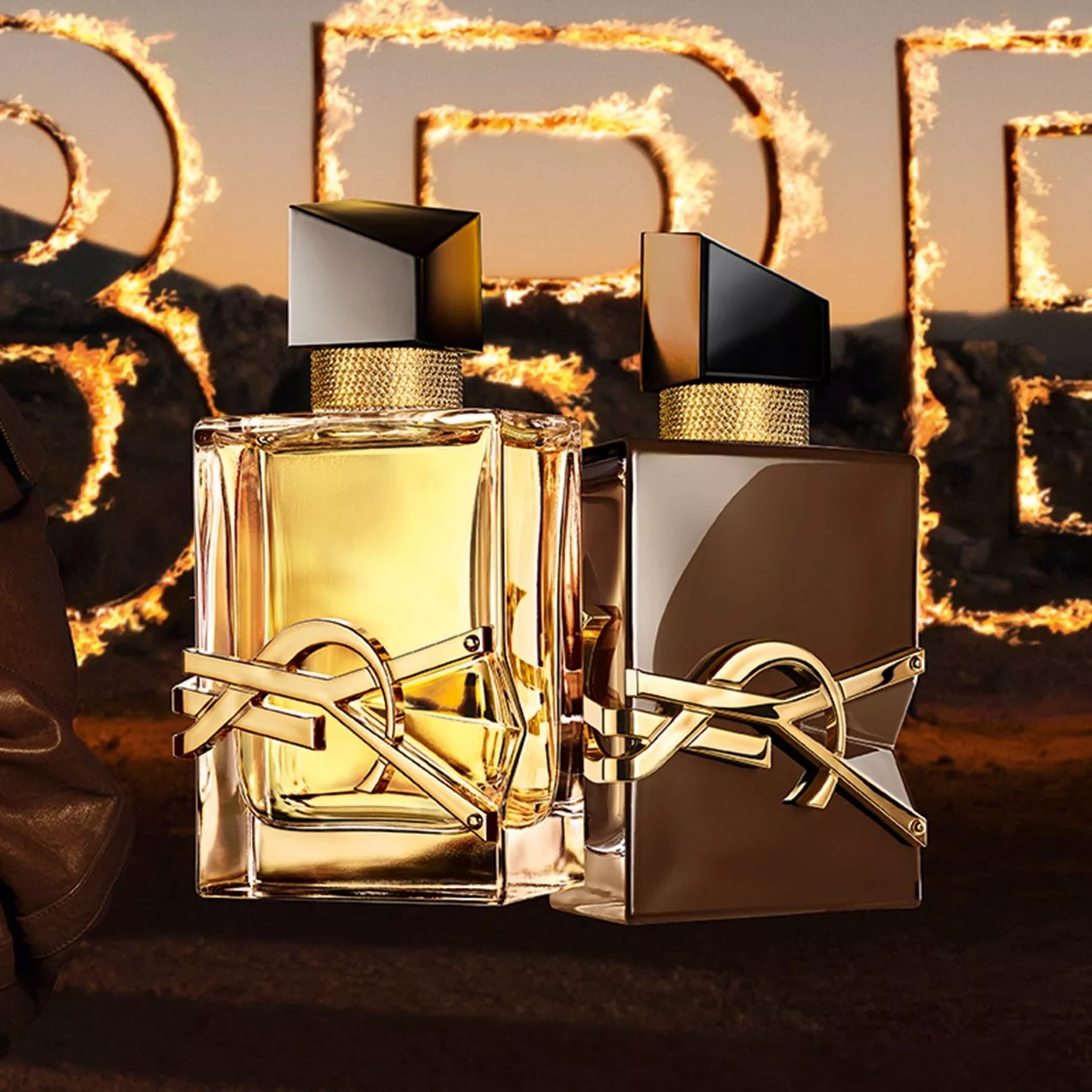 Two YSL Libre perfume bottles—one clear yellow, one dark brown—with gold logos, set against giant flaming letters in a desert sunset backdrop.