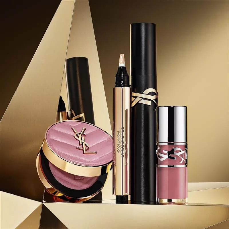 four Yves Saint Laurent makeup products—a pink quilted compact, a Touche Éclat Radiant Touch pen, a black mascara, and a pink lip gloss—arranged on reflective gold surfaces