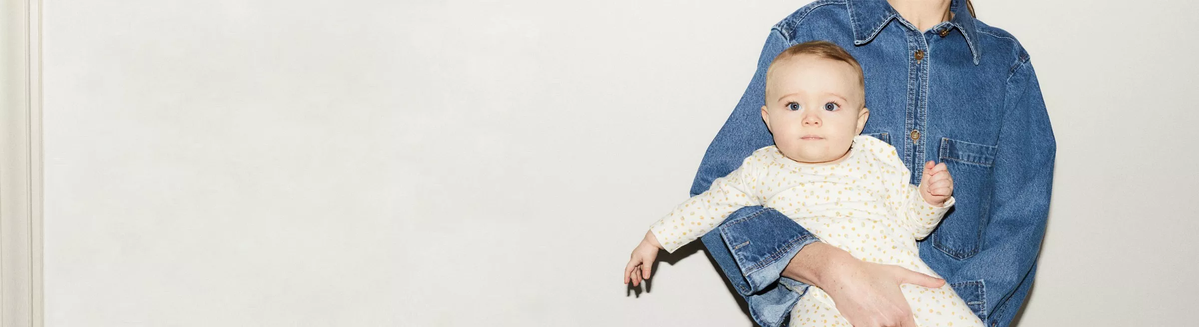 Person in denim shirt holding a baby dressed in a polka-dot outfit against a plain white background.