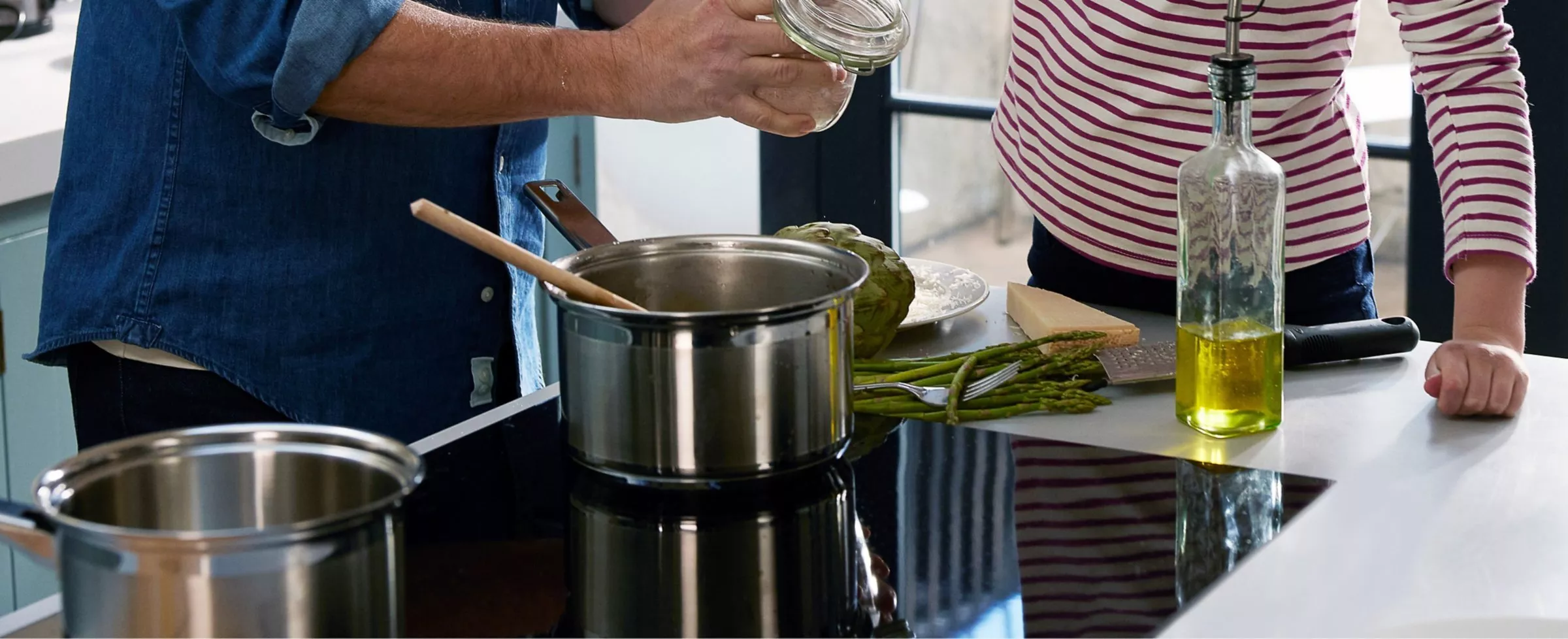 A person in a kitchen is preparing food with pots, asparagus, and olive oil on a counter.
