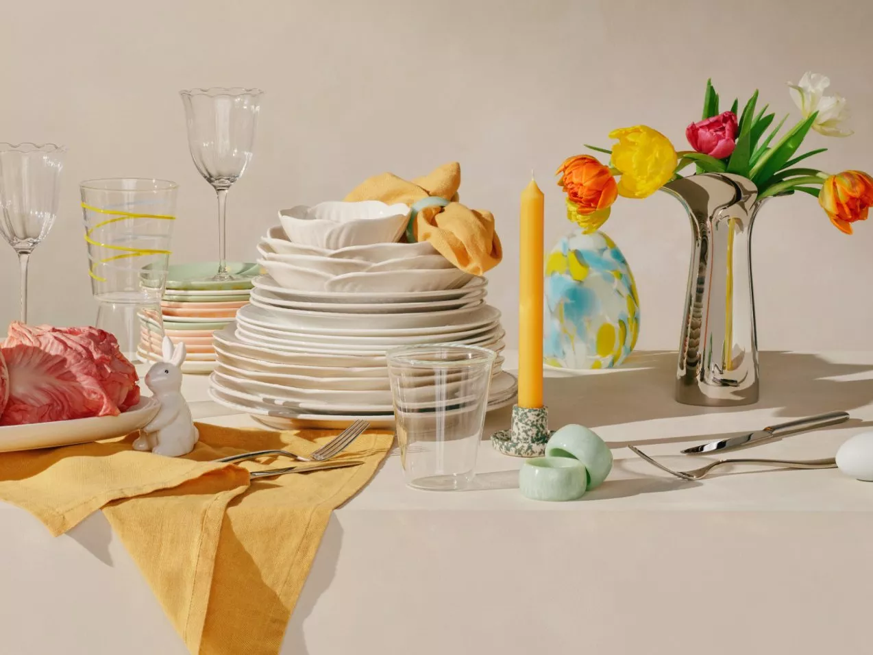 Elegant table setting with stacked plates, vibrant tulips in a vase, glassware, and a yellow candle on a cloth.