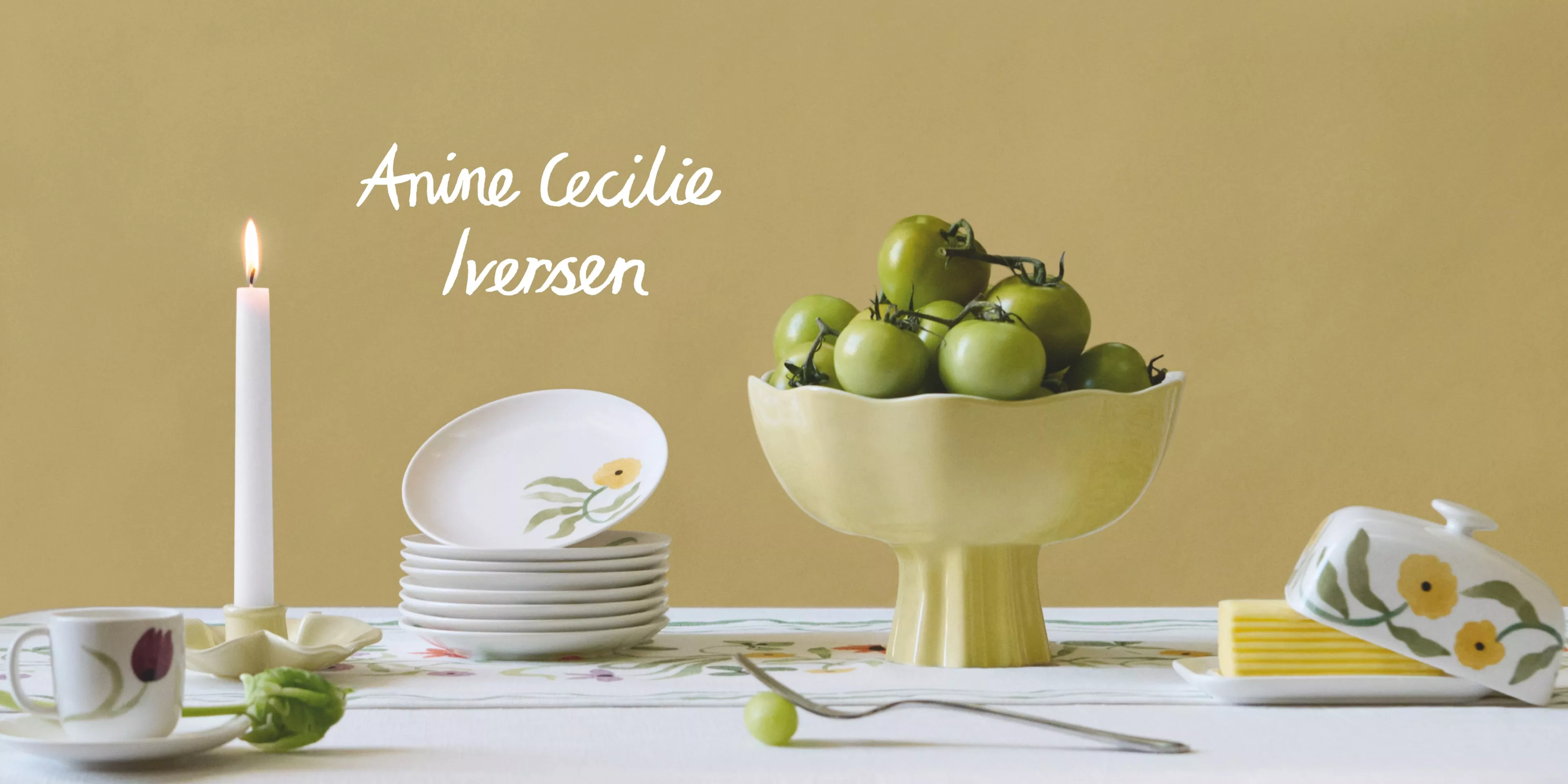 A serene table setting with green tomatoes in a bowl, floral plates, and a lit candle against a soft background.
