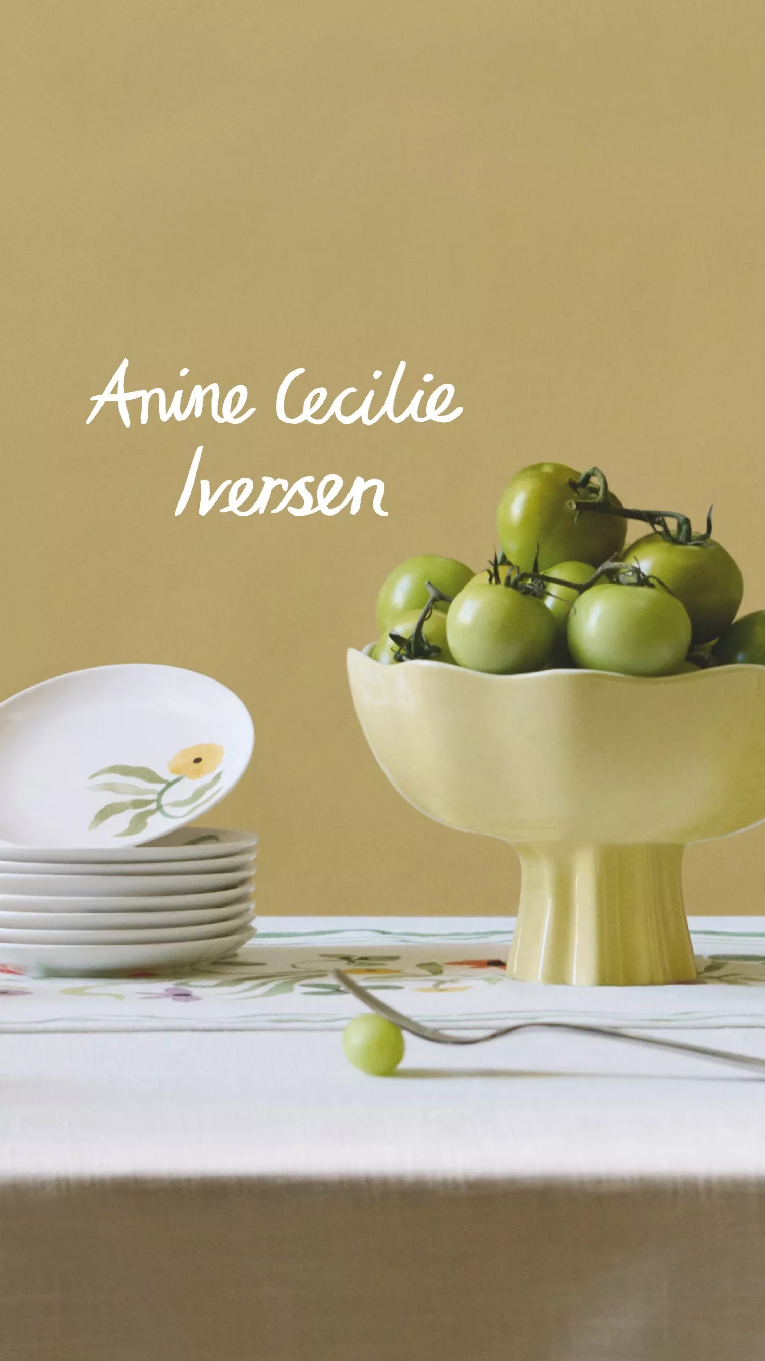 A stack of white plates with floral designs beside a yellow bowl of green tomatoes on a white tablecloth.