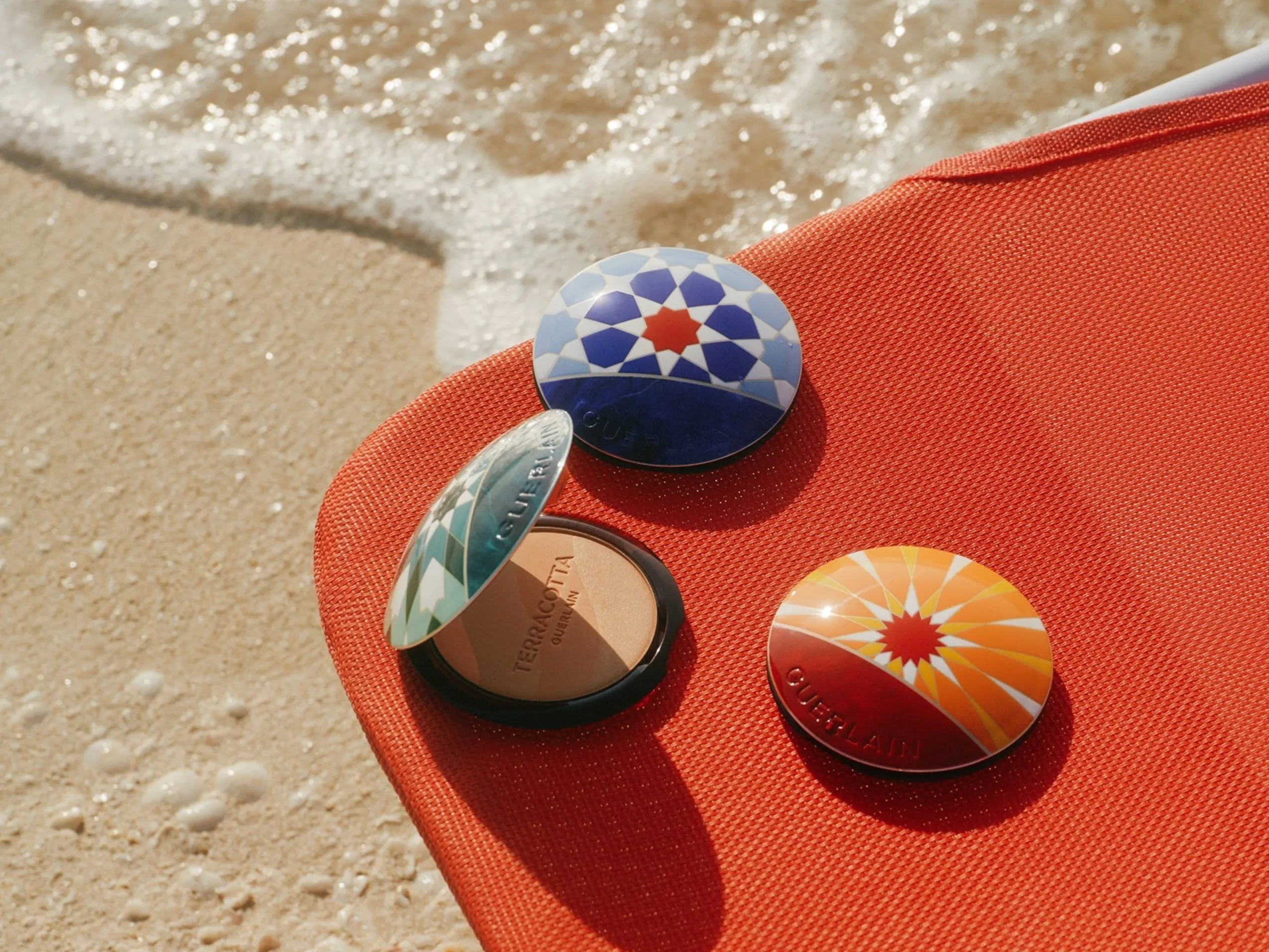 Guerlain Terracotta bronzing powder compacts on red sun lounger by the sea, with waves and sand background
