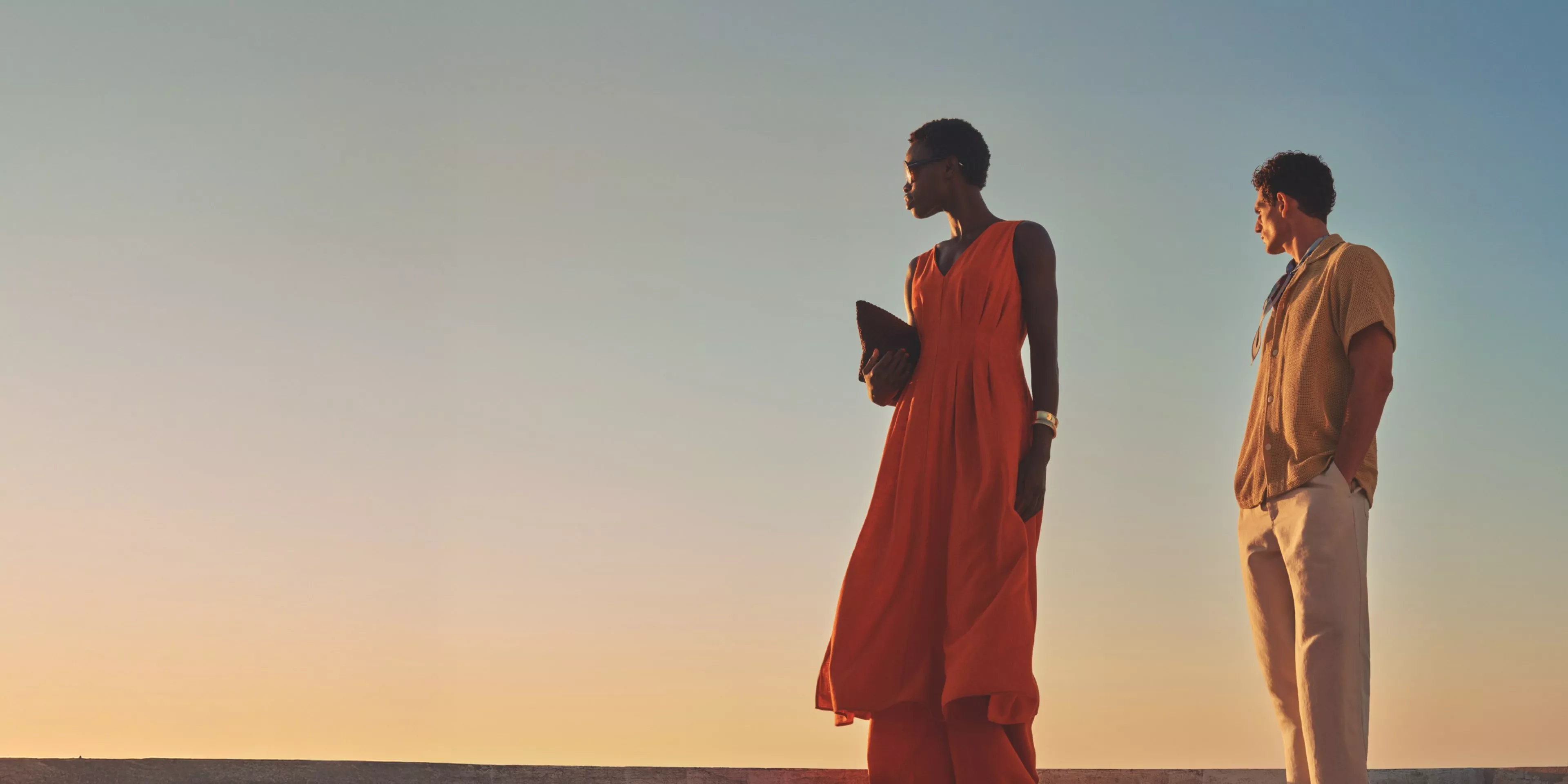 Two stylish adults in summer outfits at sunset, woman in orange maxi dress holding clutch, clear sky backdrop