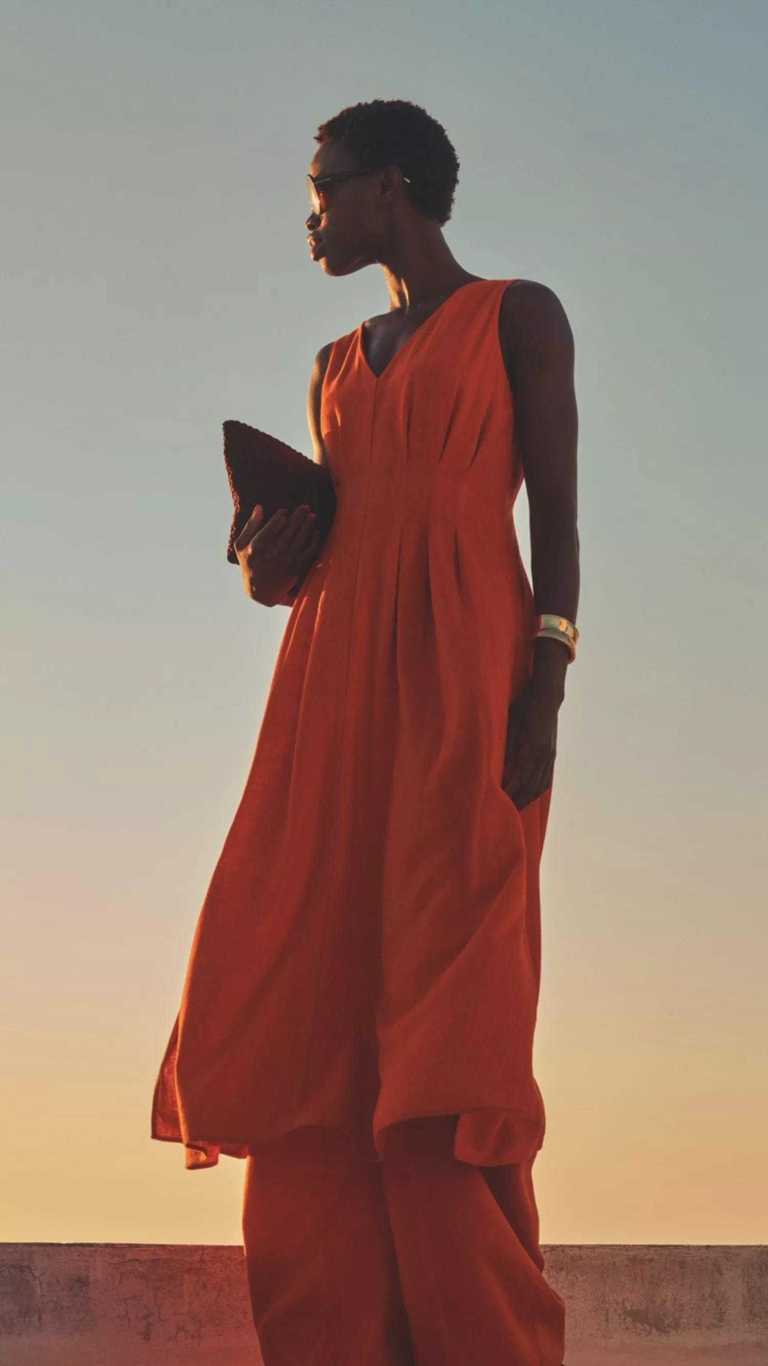 Stylish woman in orange maxi dress holding clutch at sunset