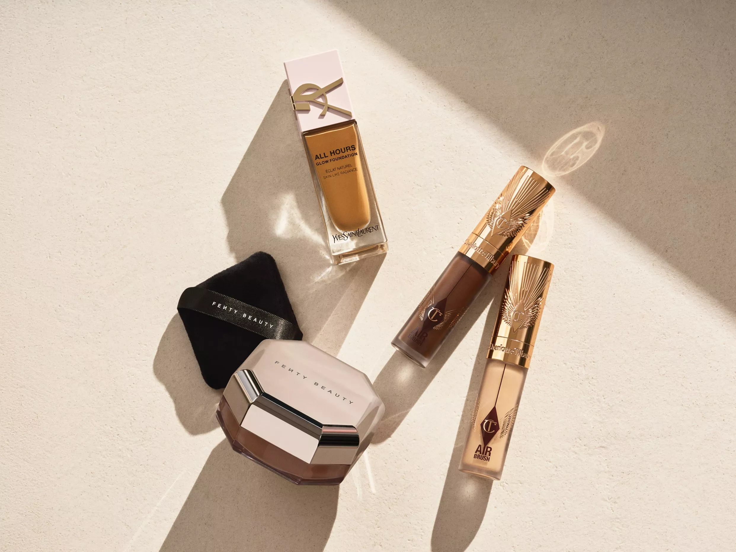 Assorted makeup products including foundation and powders are arranged on a light surface, casting soft shadows.