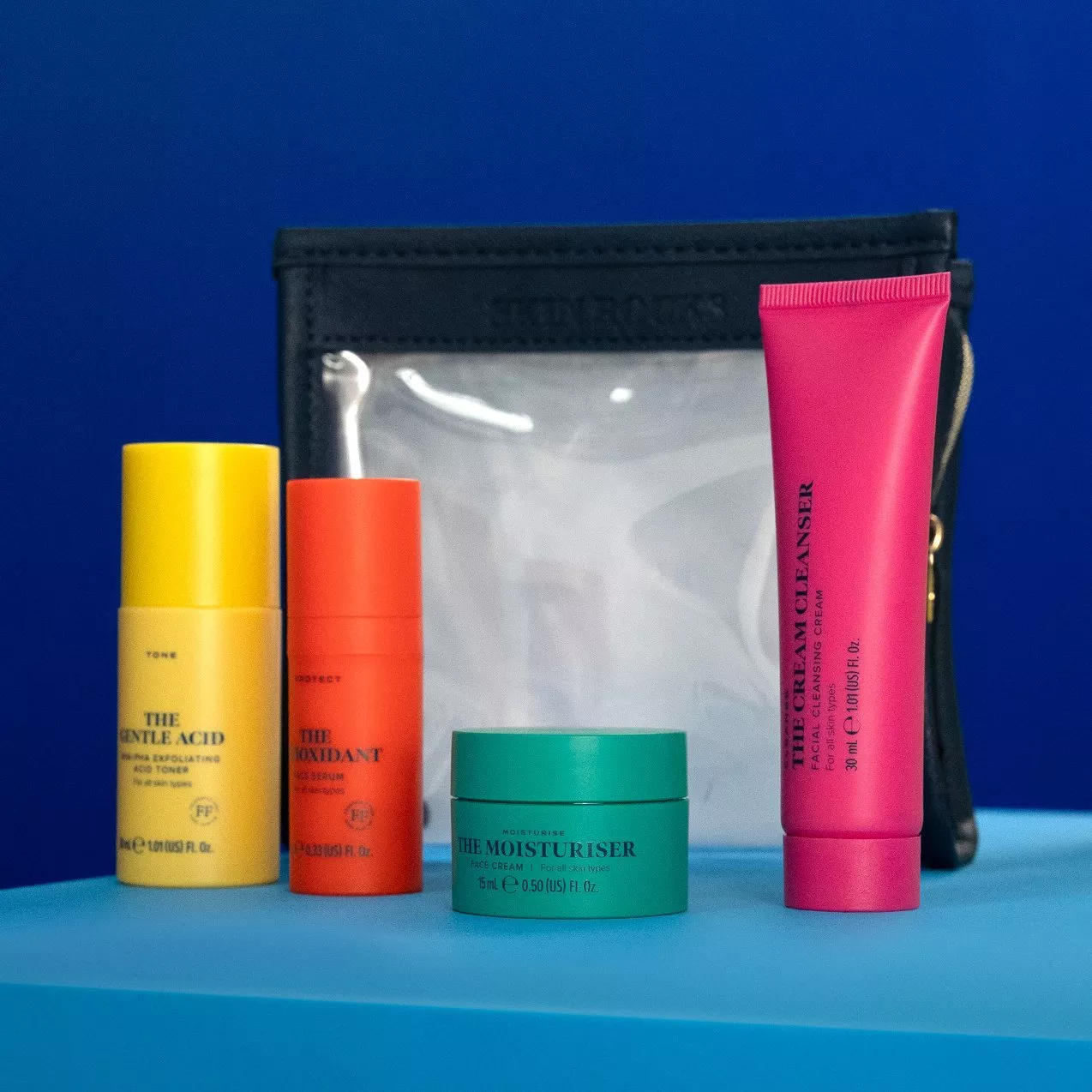 Travel skincare set with cleanser, moisturiser, toner and serum in front of a clear toiletry bag on blue background