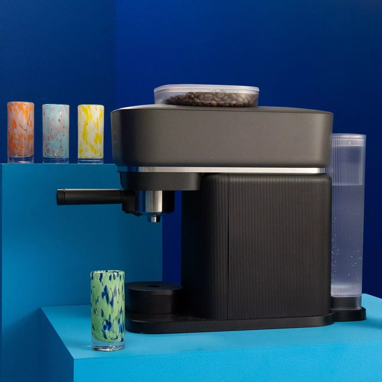 Modern black espresso coffee machine with coffee beans hopper and water tank on blue counter, colourful glasses nearby