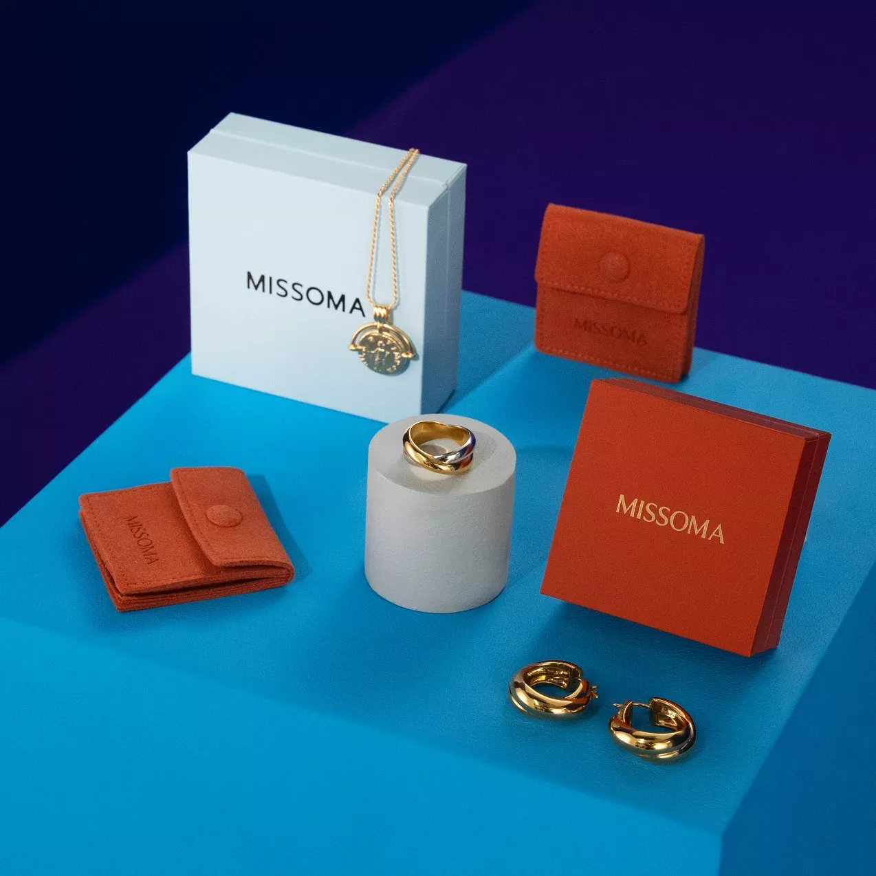 Missoma jewellery gift set with gold necklace, ring and hoop earrings, plus branded boxes and pouches on blue plinth.