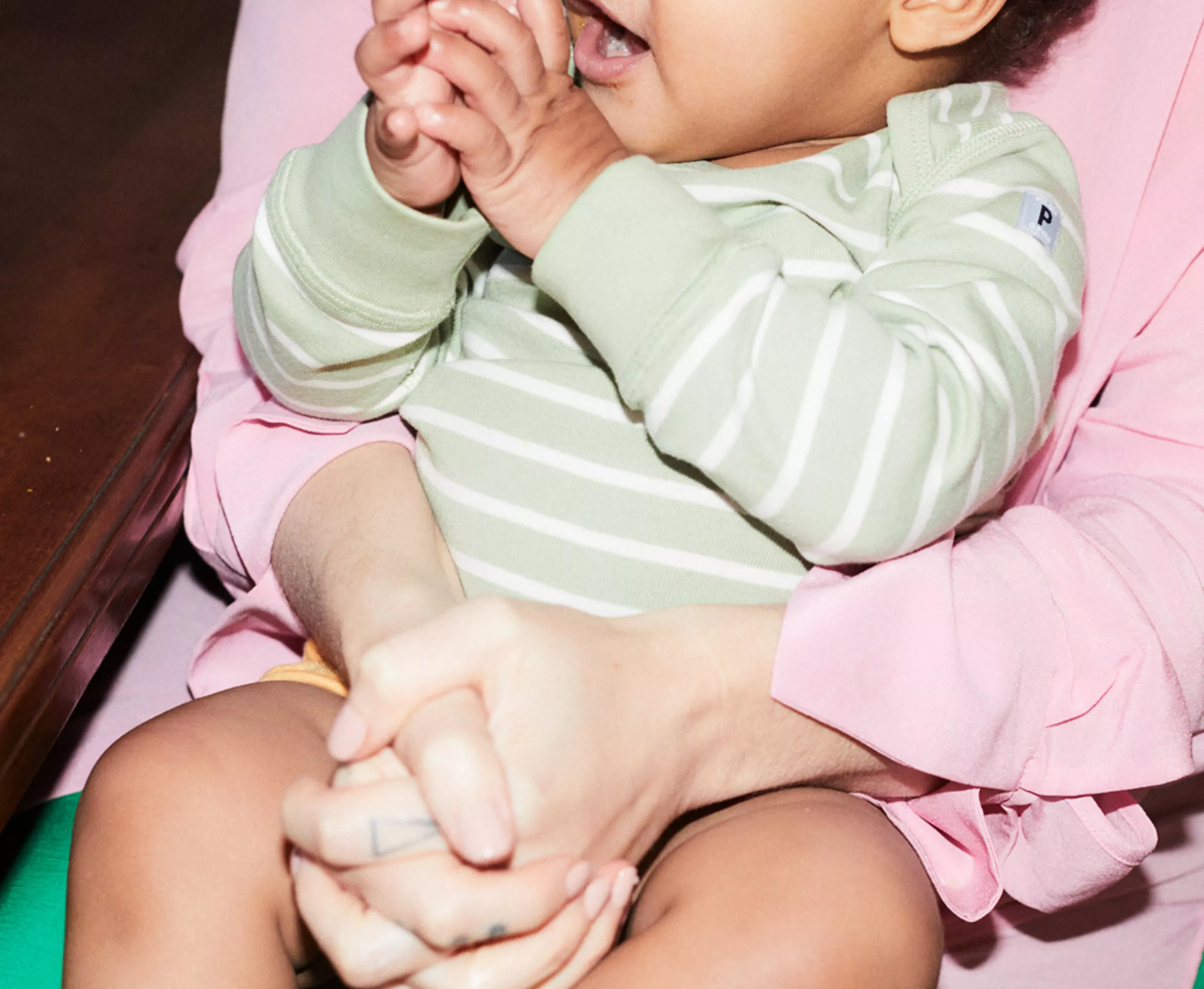 A baby in a green striped outfit claps hands while sitting on an adult's lap