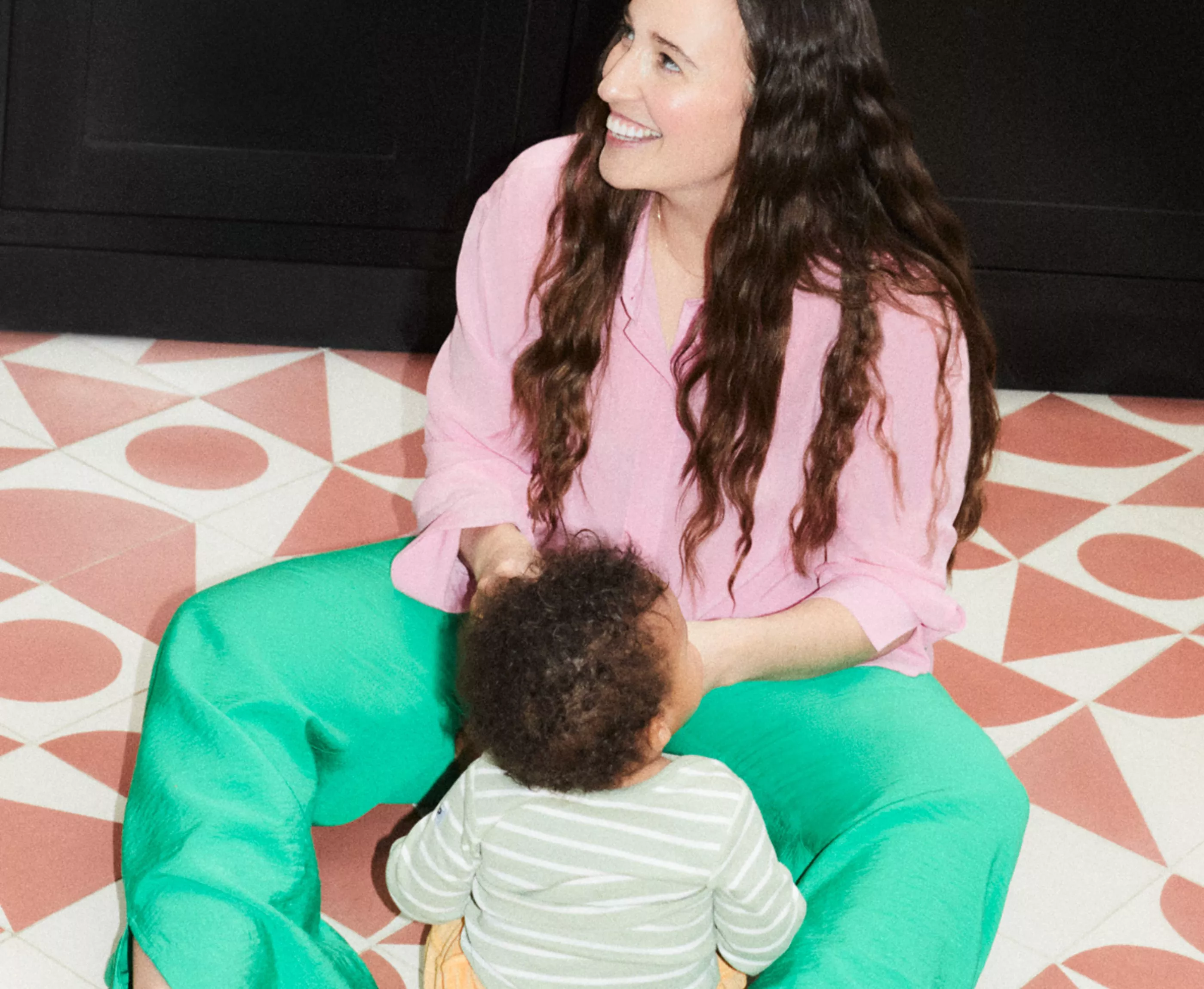 A woman in a pink blouse and green trousers sits on patterned tiles, smiling at a child in front of her.