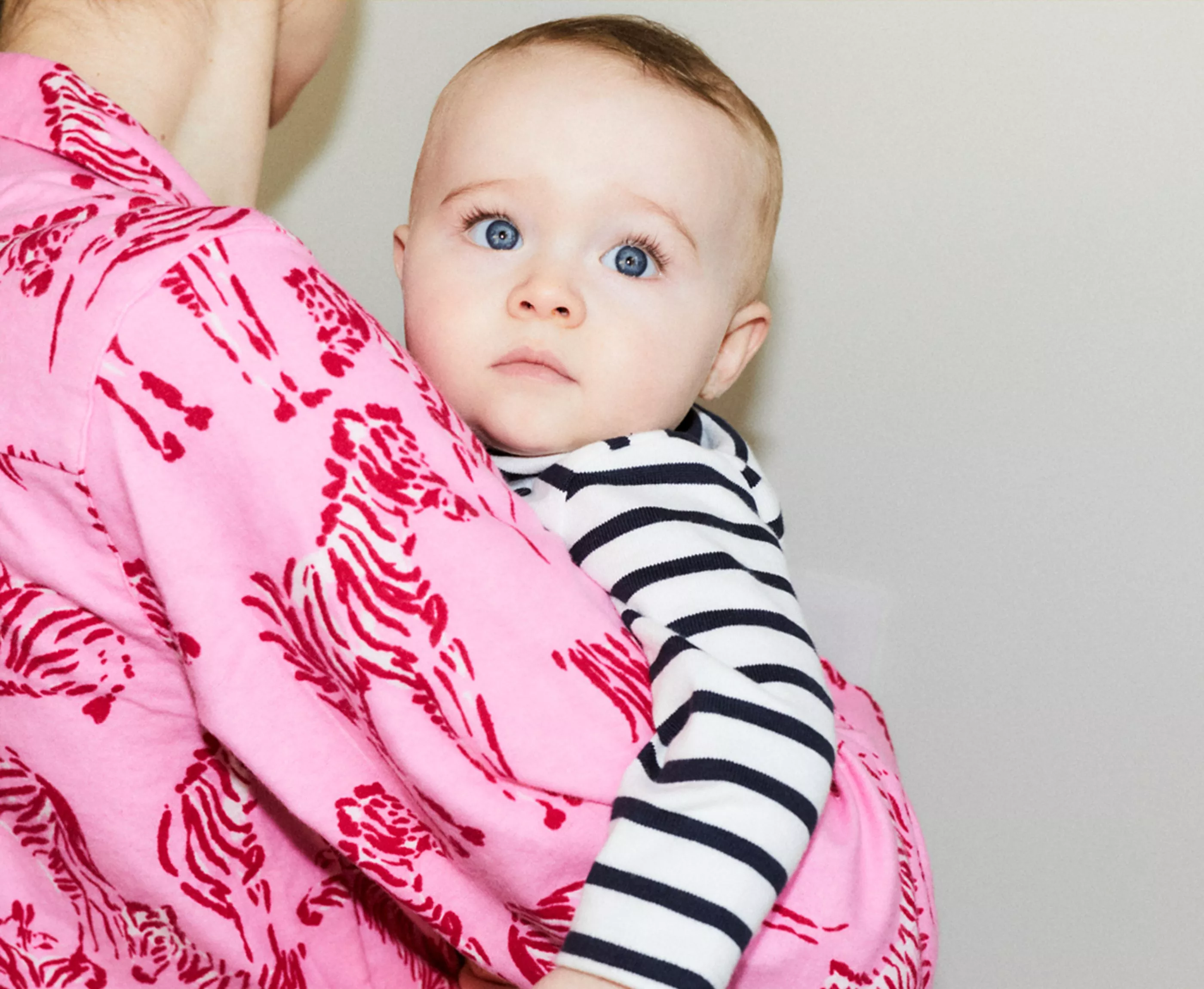 A baby in a striped outfit is held by someone in a pink, zebra-patterned jumper