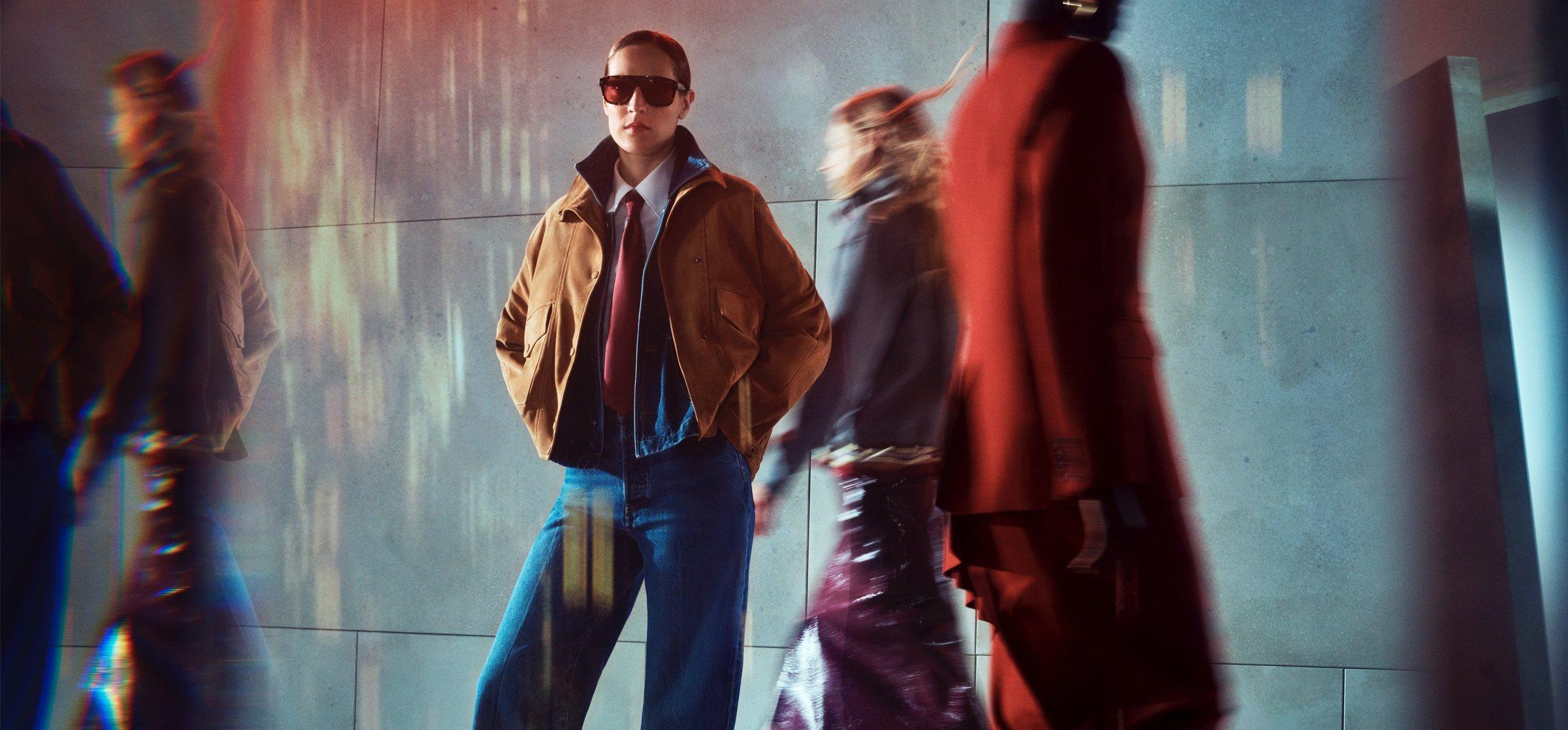 A female model in an autumn look with a tan leather jacket, pausing with sunglasses on and a shirt and tie